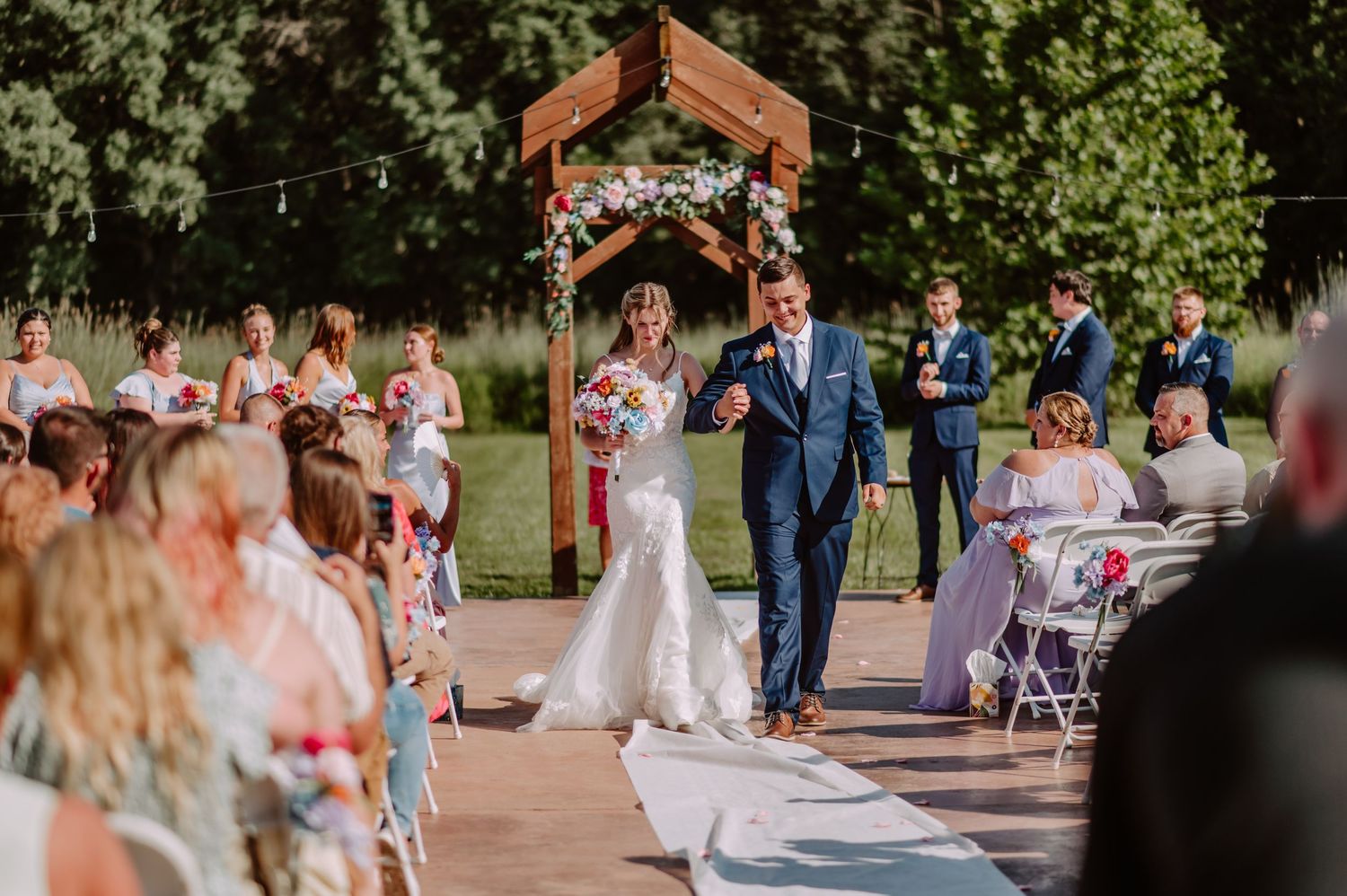 Bride and father walk down aisle at outdoor wedding ceremony with guests seated on sides.
