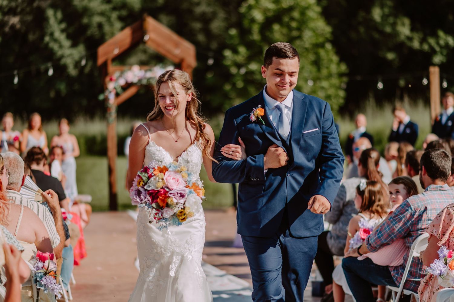 Beaming bride with vibrant bouquet and groom in navy suit joyfully walk outdoor wedding ceremony aisle.