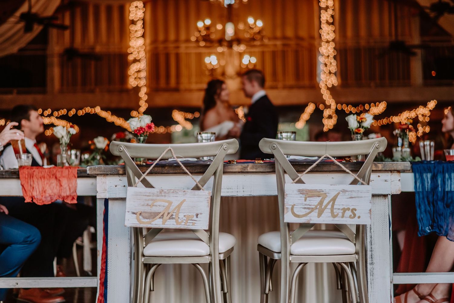Rustic barn wedding reception with Mr. and Mrs. chairs at sweetheart table, string lights, and couple embracing in background.