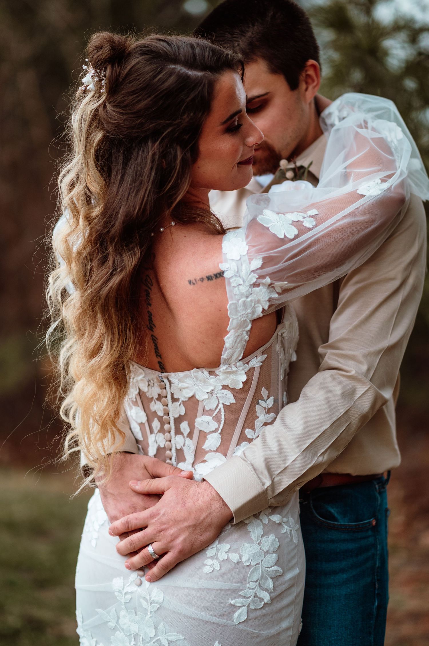 A bride in a floral lace backless gown embraces her groom outdoors in a romantic wedding photo.