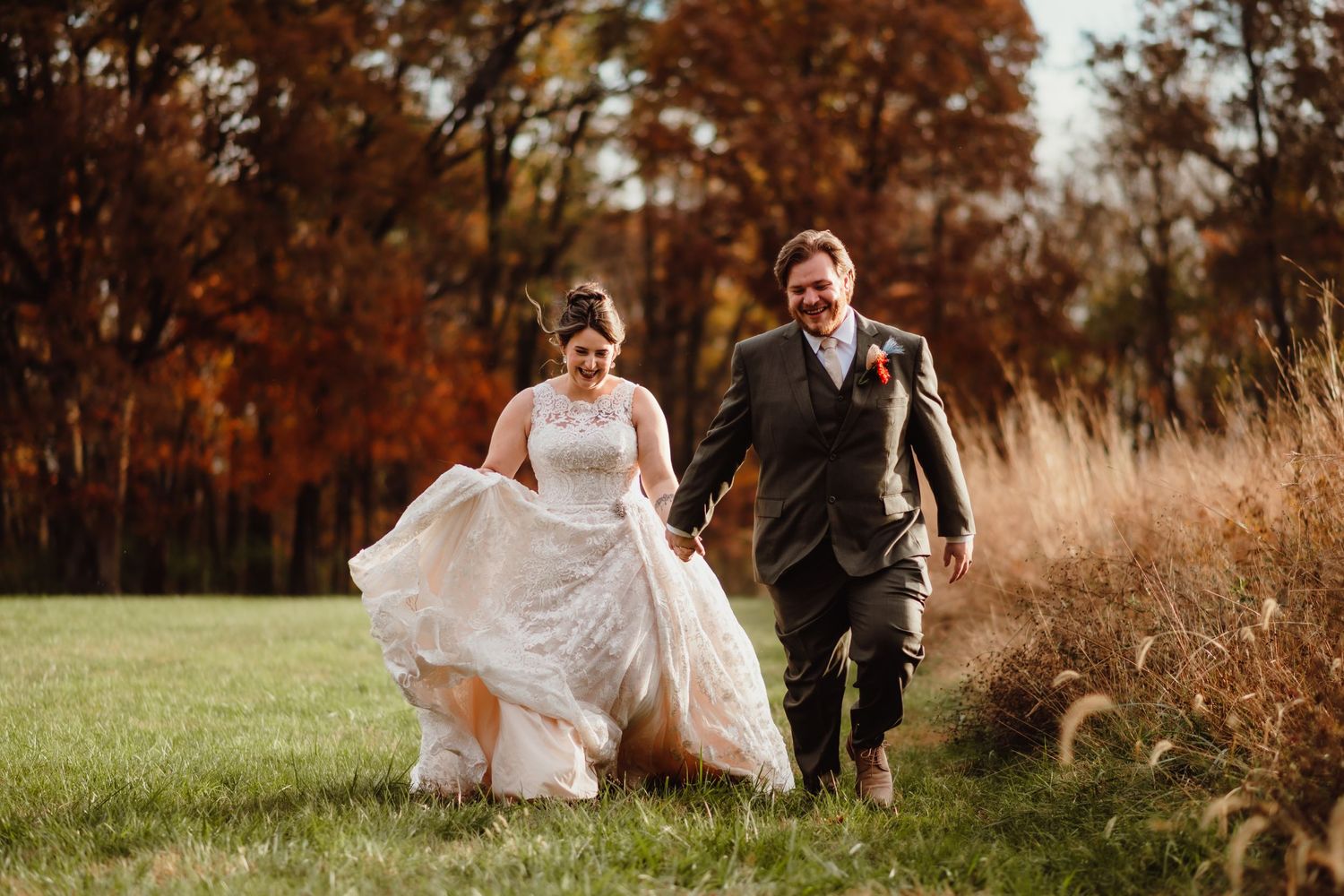 Bride and groom laughing and running through a green field with vibrant autumn foliage in the background.