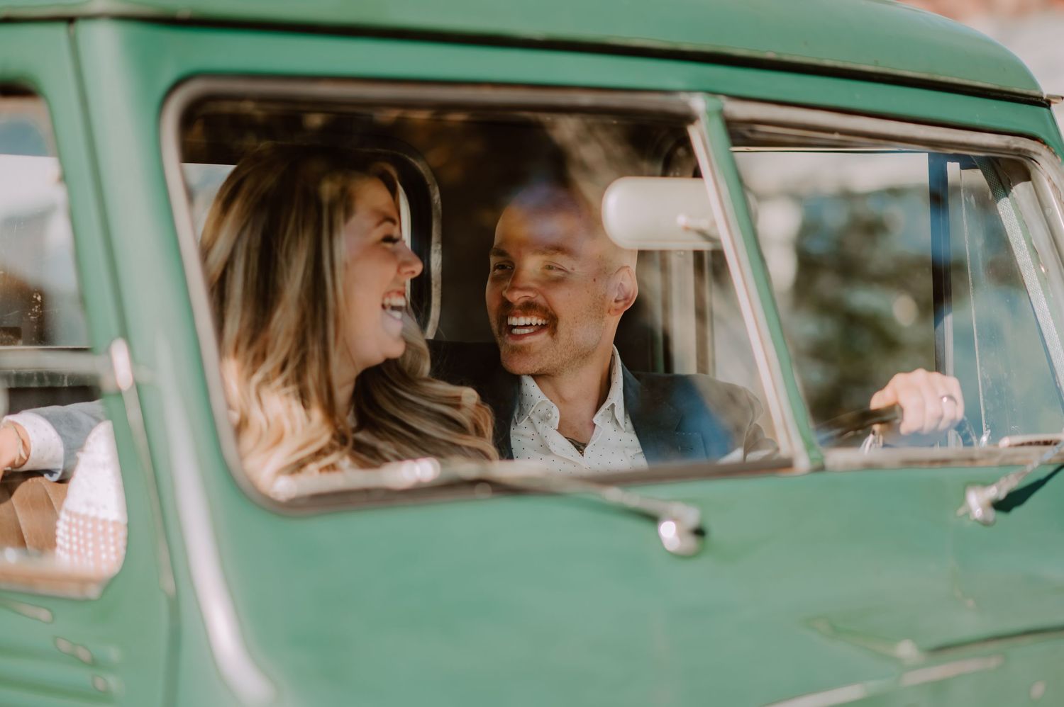 Happy couple laughing together inside a vintage teal VW bus, sharing a joyful moment on a sunny day.
