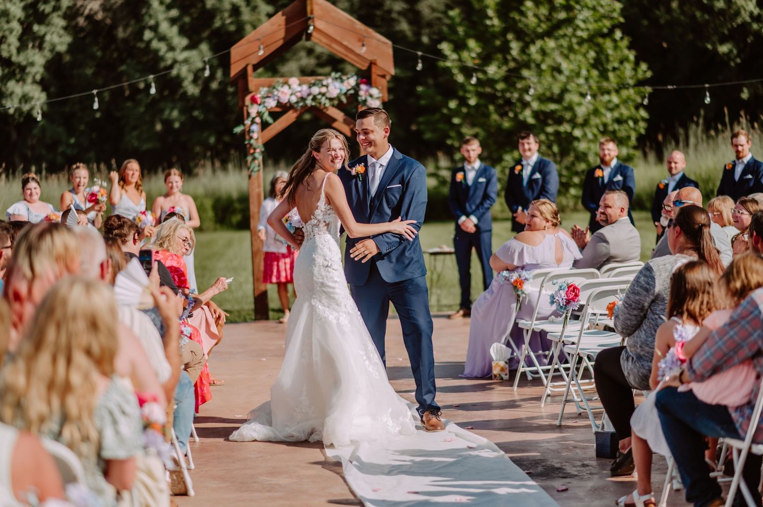 Smiling bride and groom walk down aisle at sunny outdoor wedding ceremony with guests.