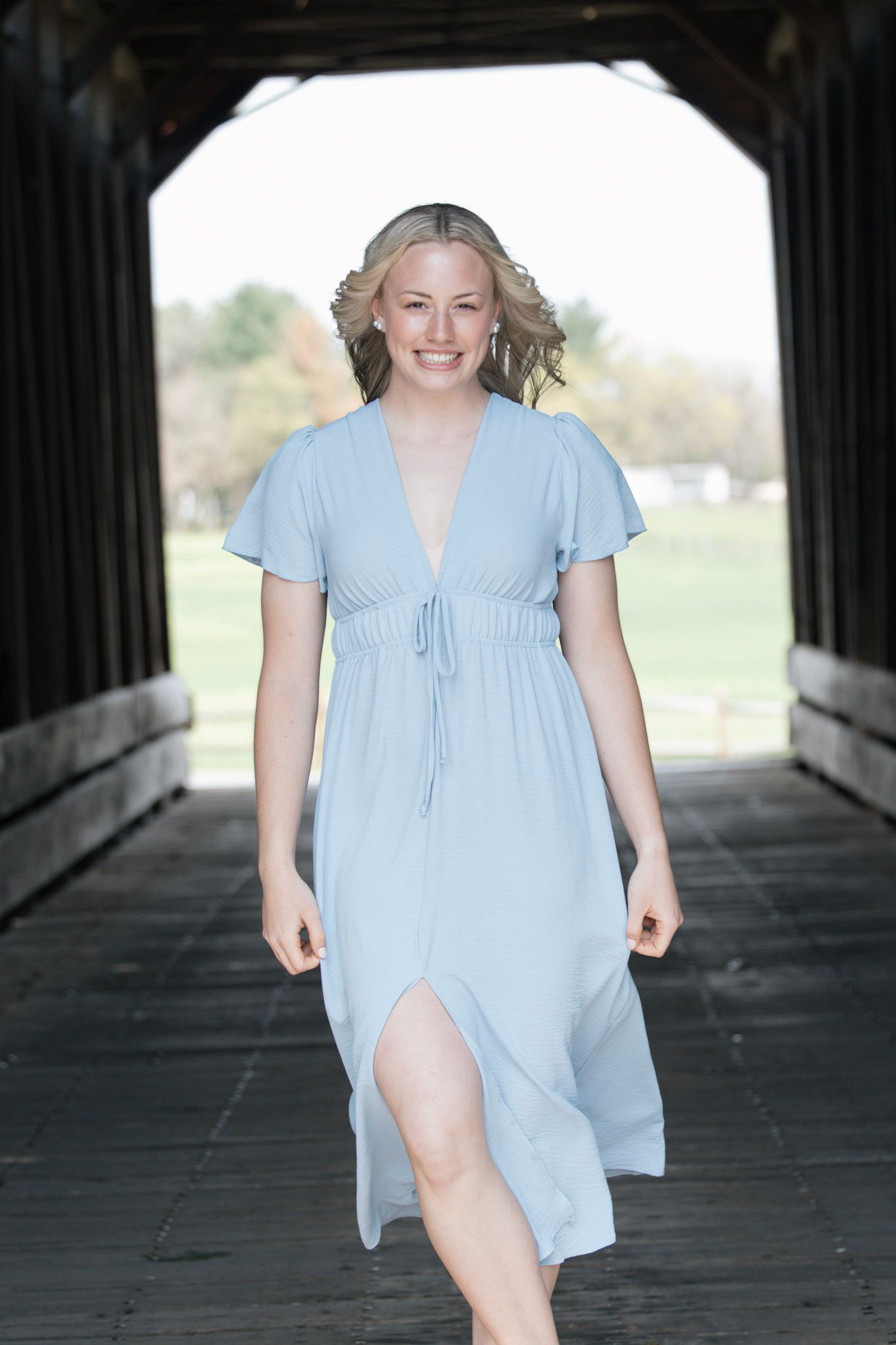Smiling blonde woman in light blue flutter sleeve dress walking through covered wooden bridge.