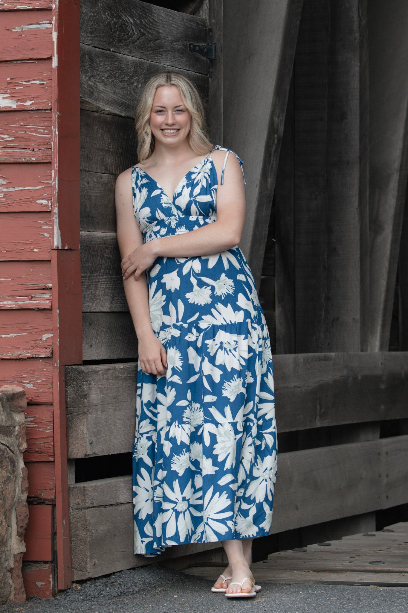 Smiling blonde woman in blue floral maxi dress standing by rustic wooden barn door.