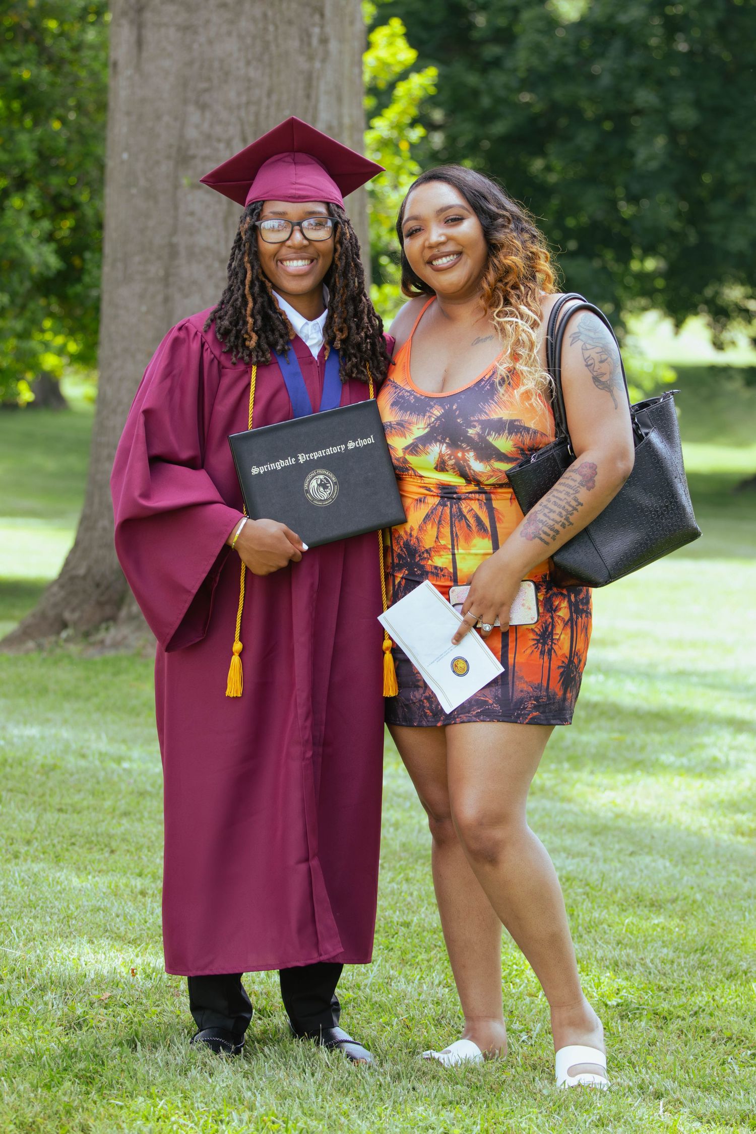 Graduate in maroon cap and gown poses with friend outdoors holding diploma at graduation ceremony.