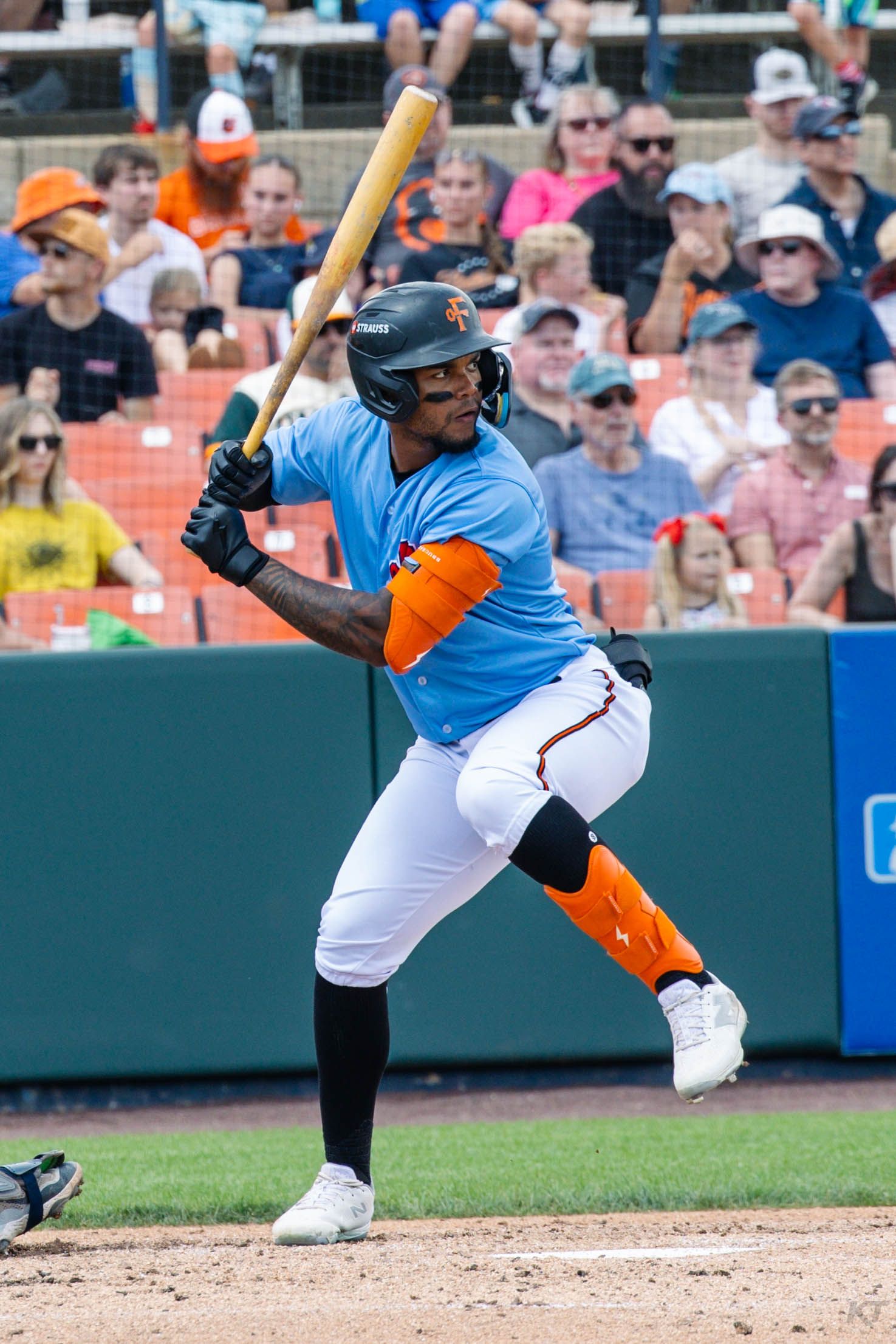 Baseball batter in blue and orange uniform swings at pitch during a game with fans watching in the stands.