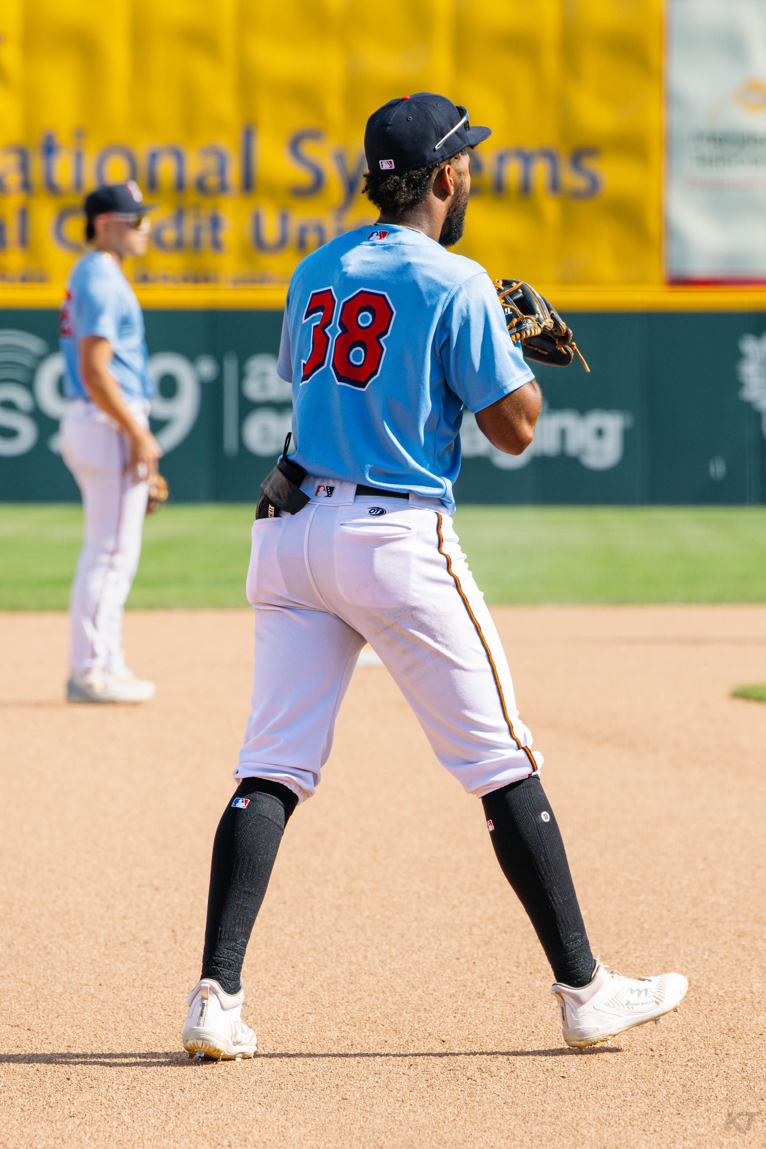 Baseball player #38 in light blue uniform stands on infield dirt during minor league game with teammate in background.