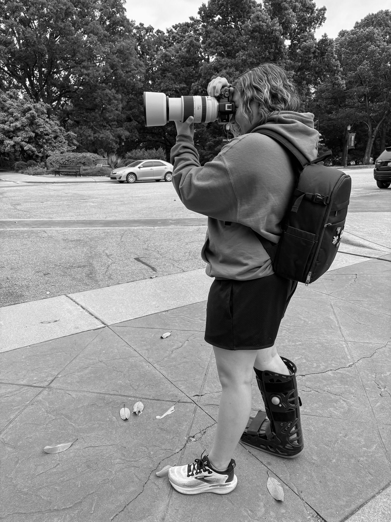 Photographer with telephoto lens, backpack, and medical boot captures outdoor shots in black and white.