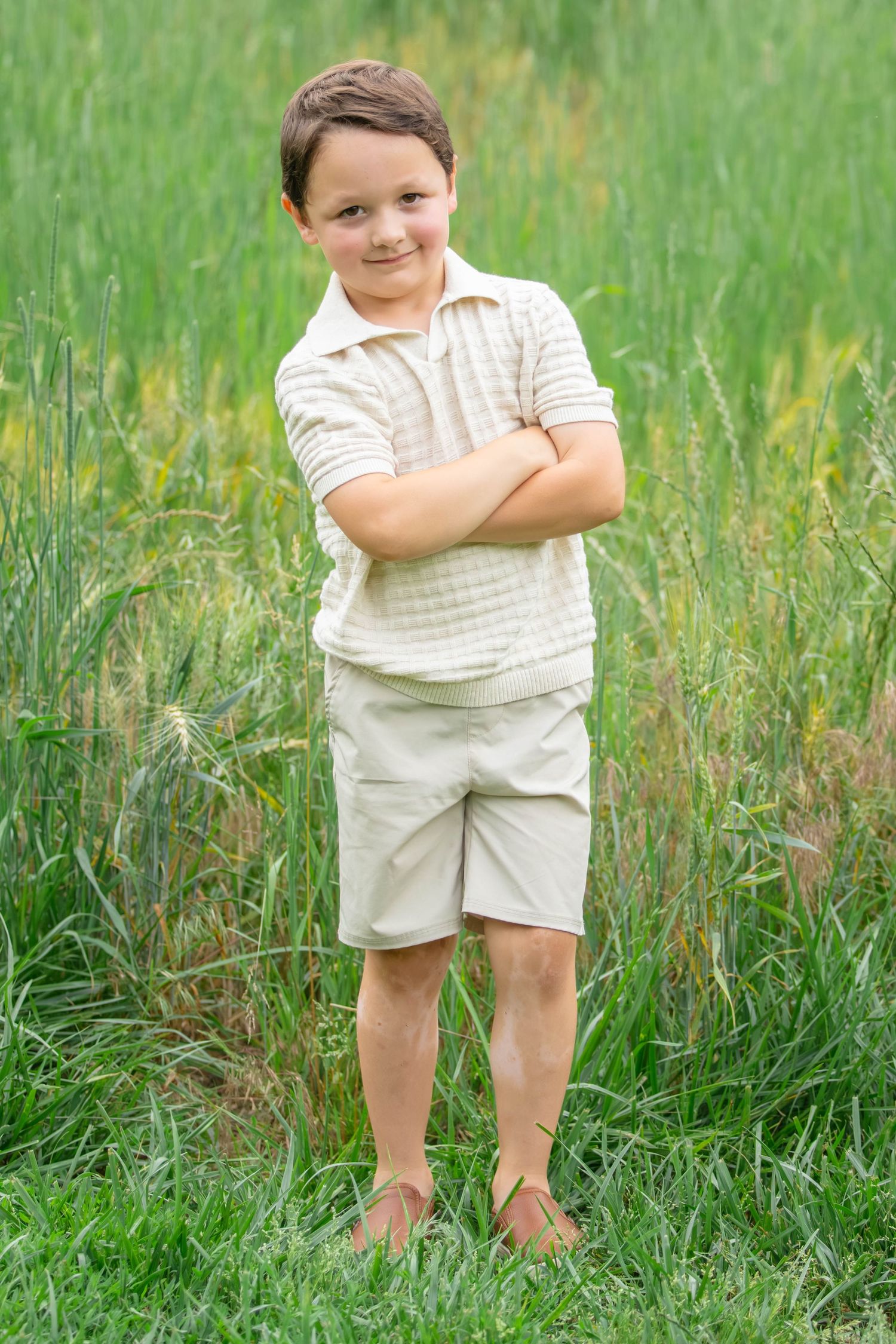 A young boy in beige shorts and polo shirt stands confidently with arms crossed in a lush green field.