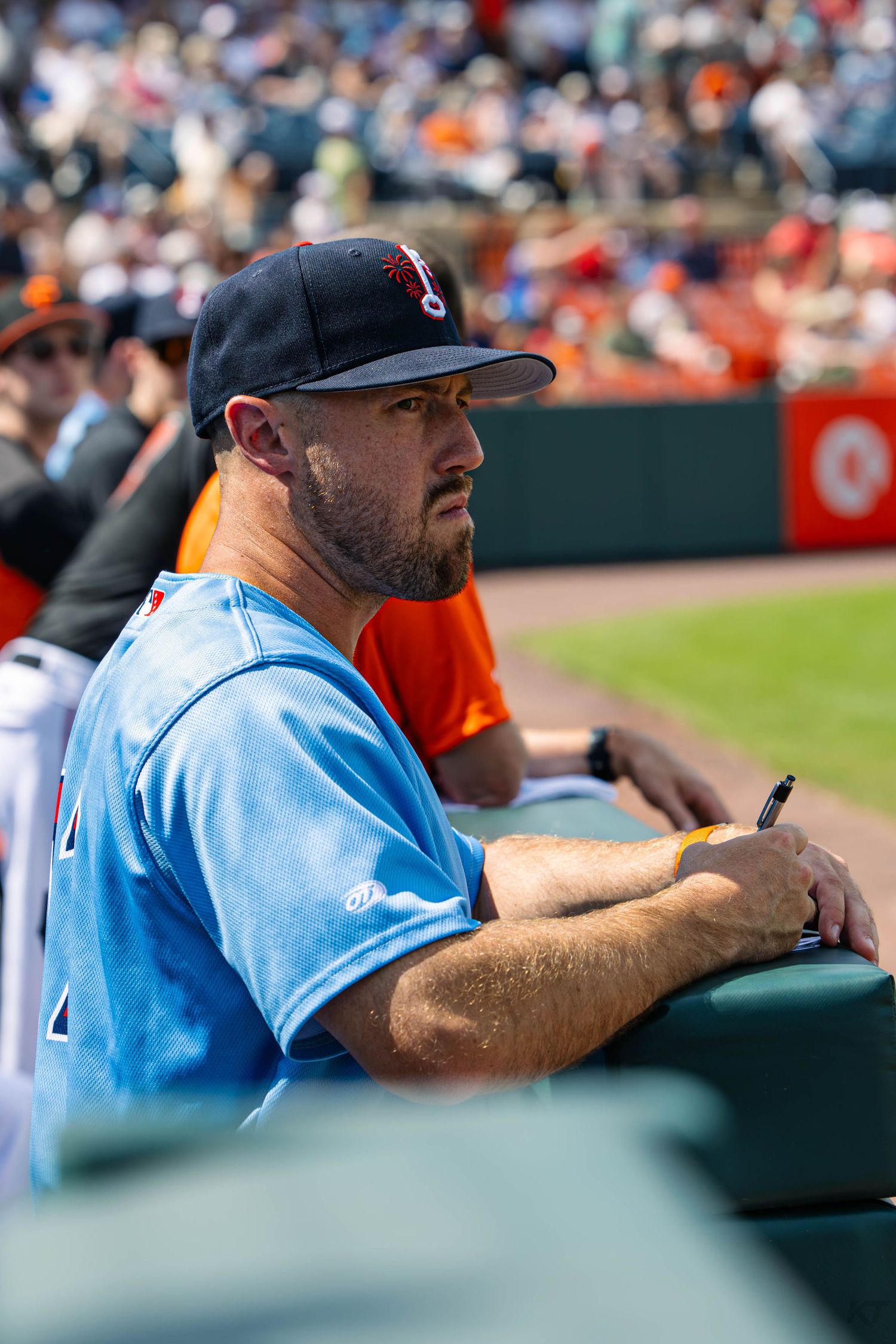 A baseball coach in a light blue jersey and black cap sits in the dugout, attentively watching the game.