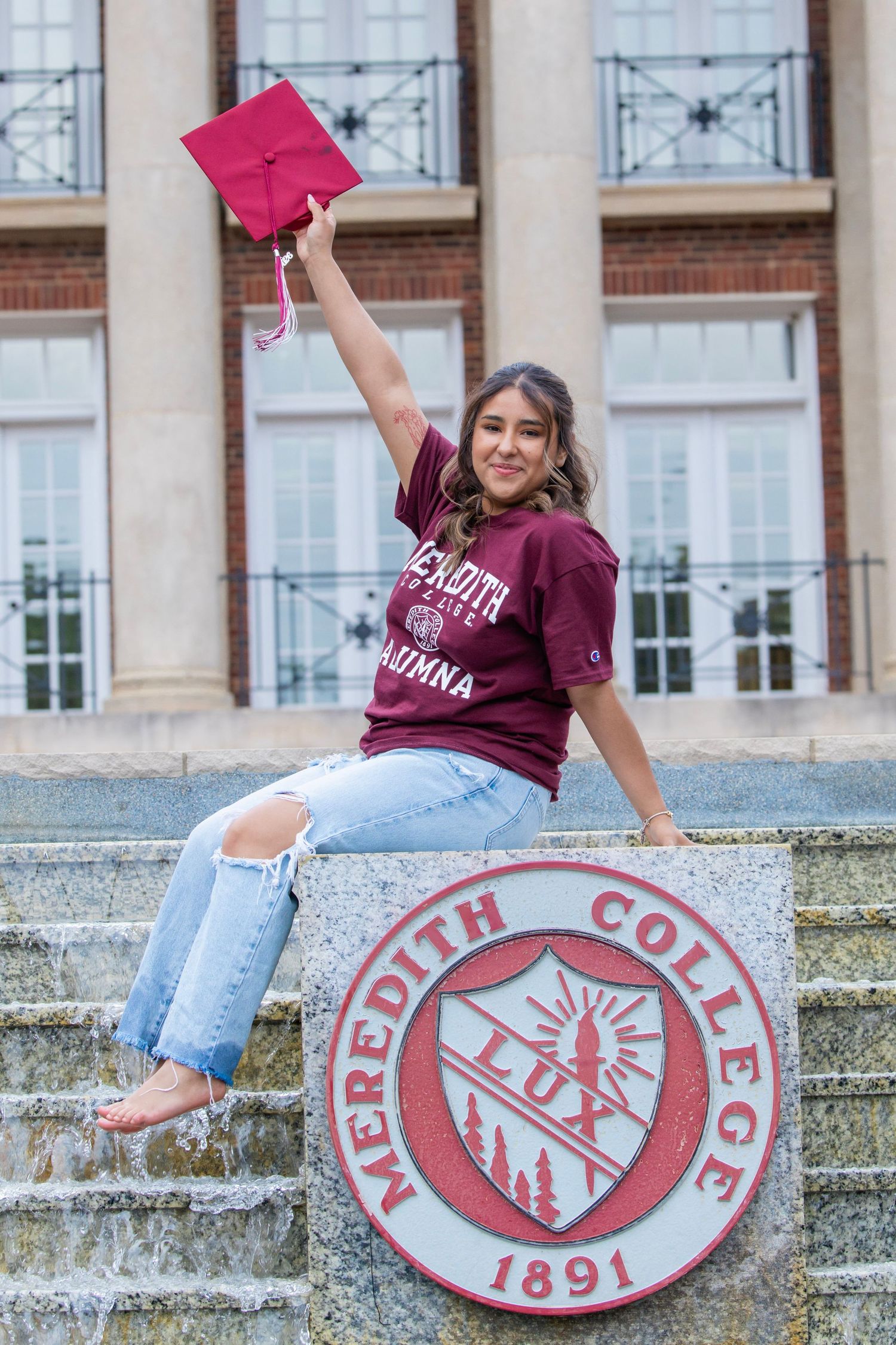 Female graduate sits on Meredith College 1891 seal fountain raising red mortarboard cap in celebration.