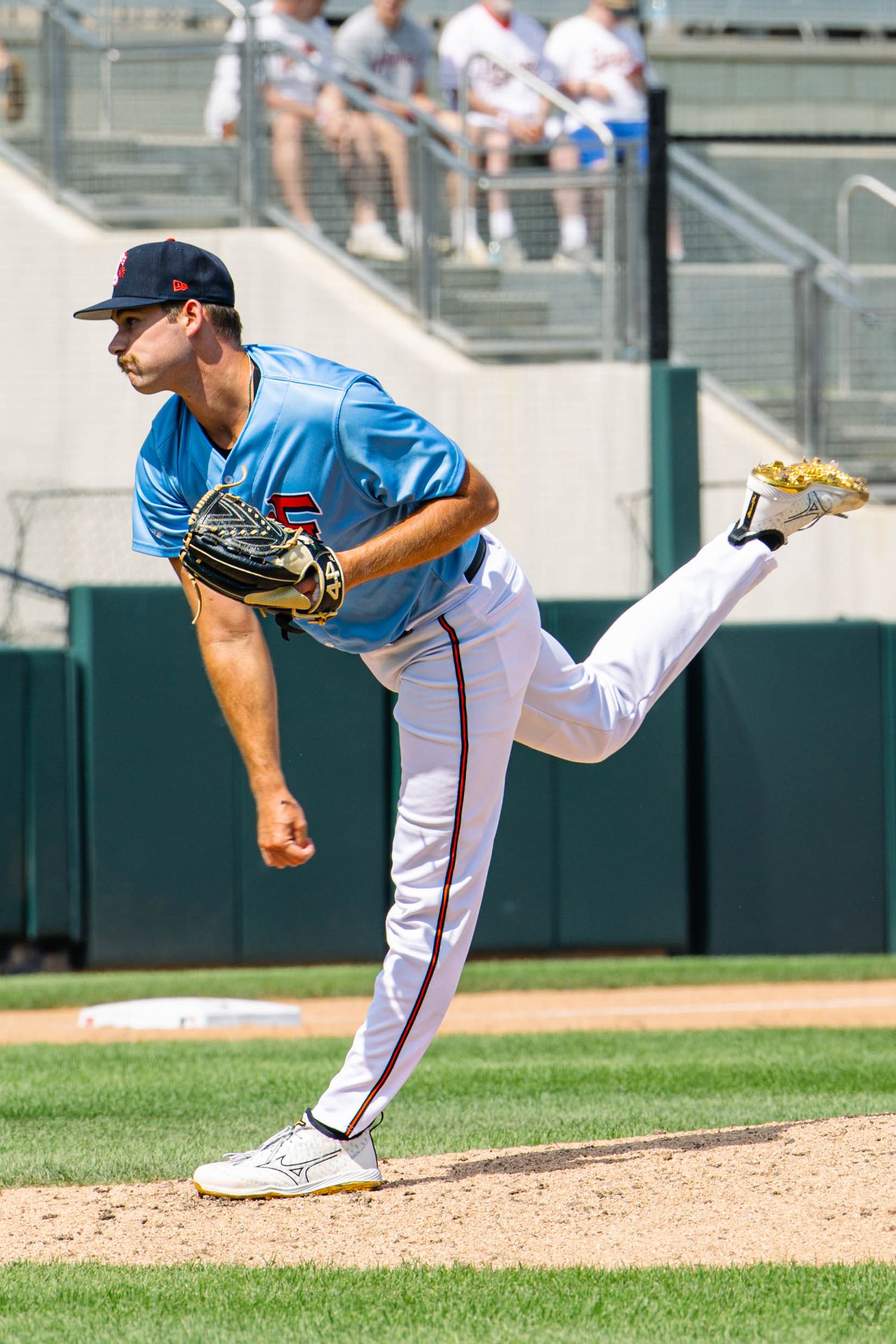 A minor league baseball pitcher in a light blue uniform delivers a pitch during a sunny afternoon game.