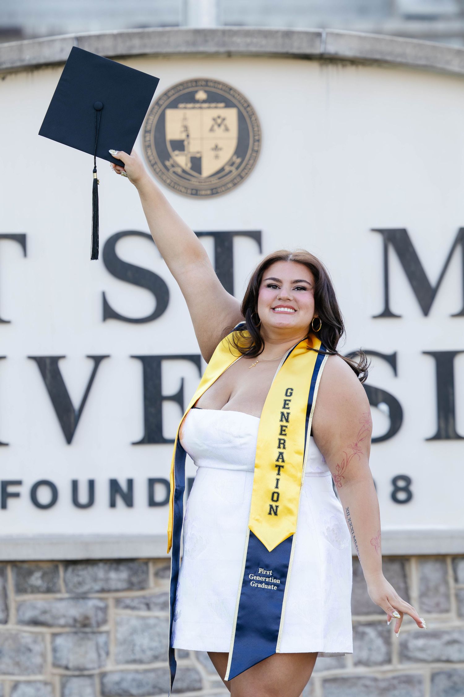 Graduate in white dress raising cap at St. Mary's University sign.