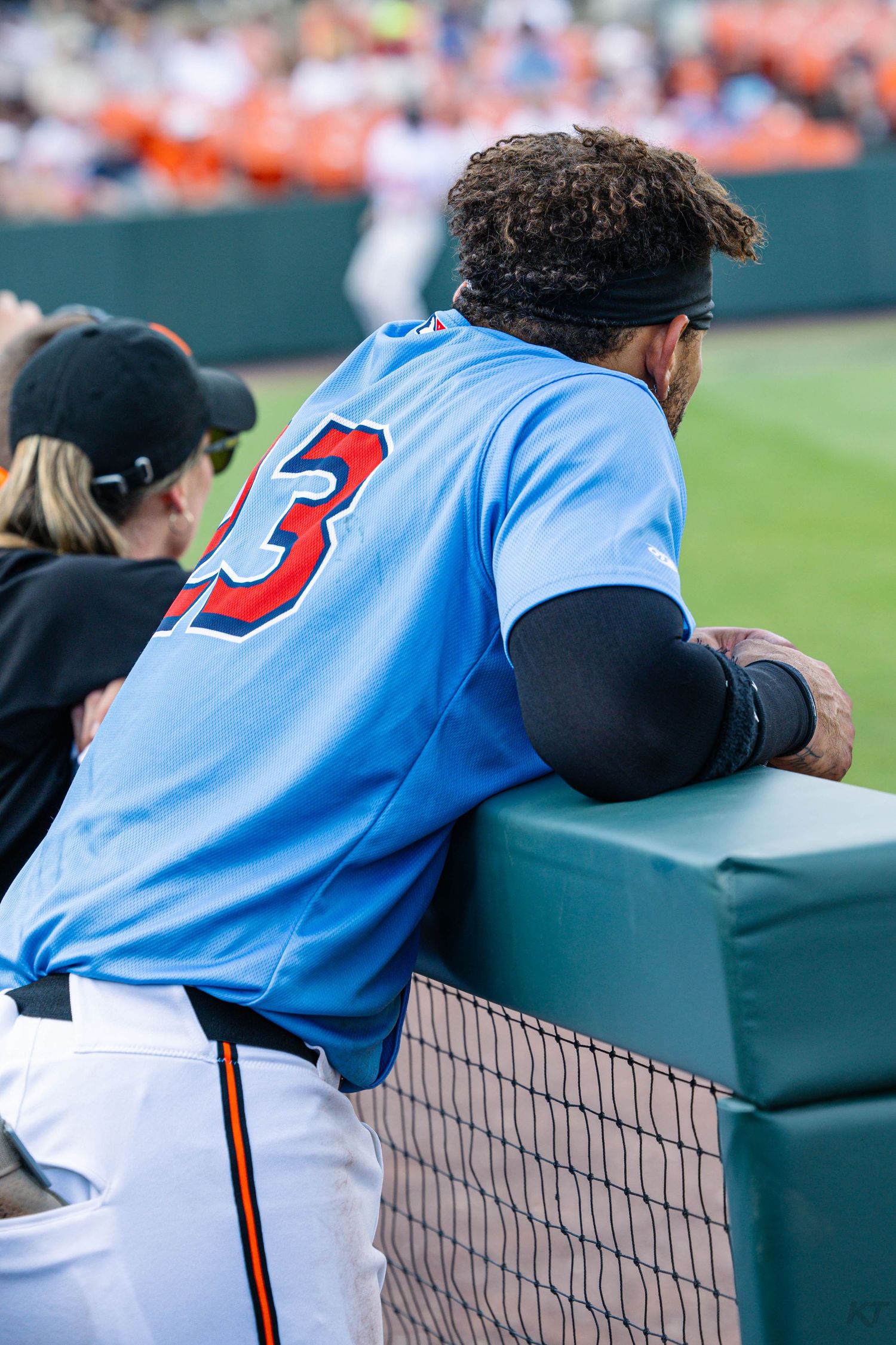 Baseball player wearing number 3 light blue jersey leans on dugout railing watching the game from the sideline.
