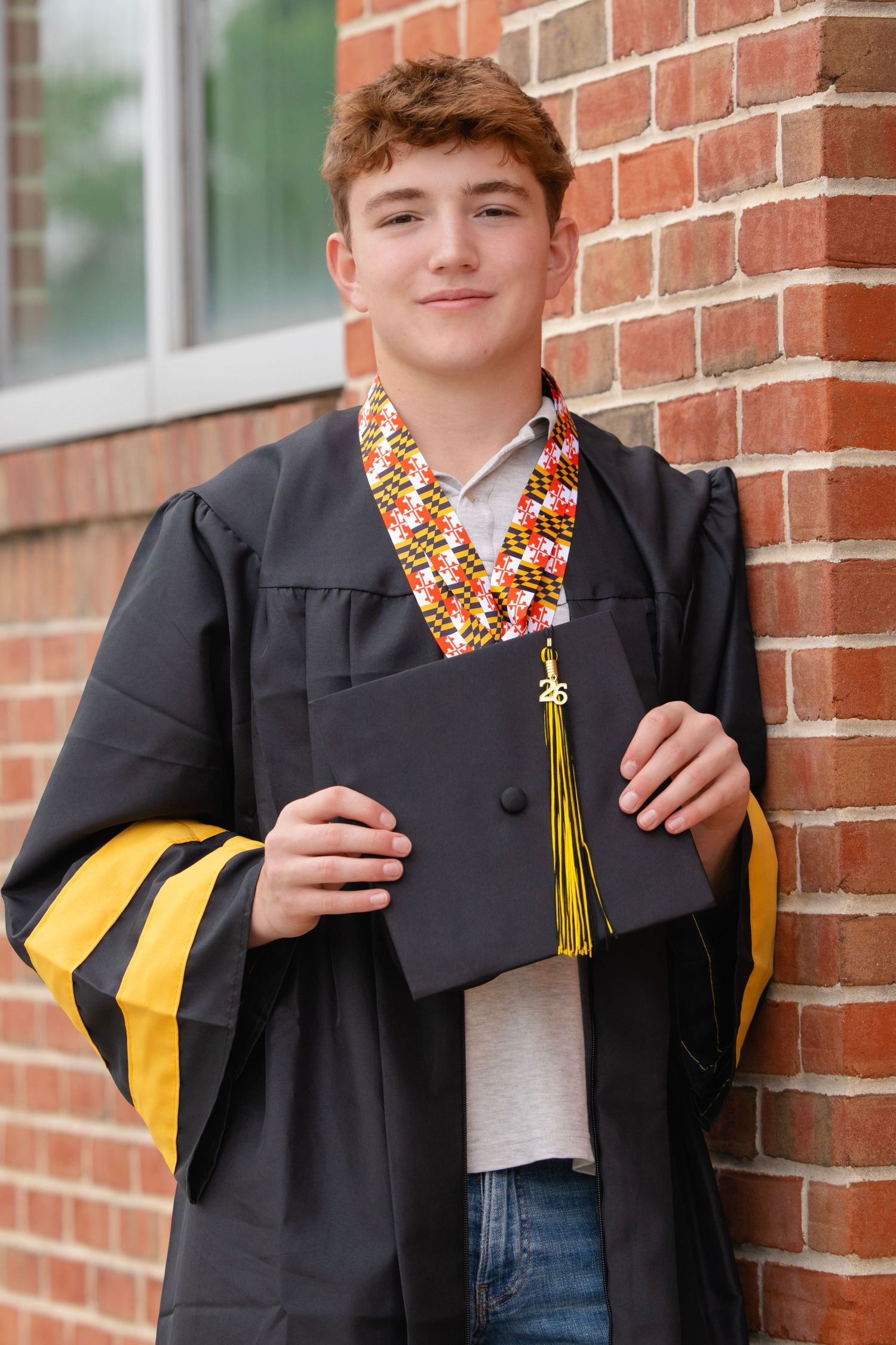 Young man in black graduation gown with gold trim and colorful stole holds cap while leaning against brick wall.