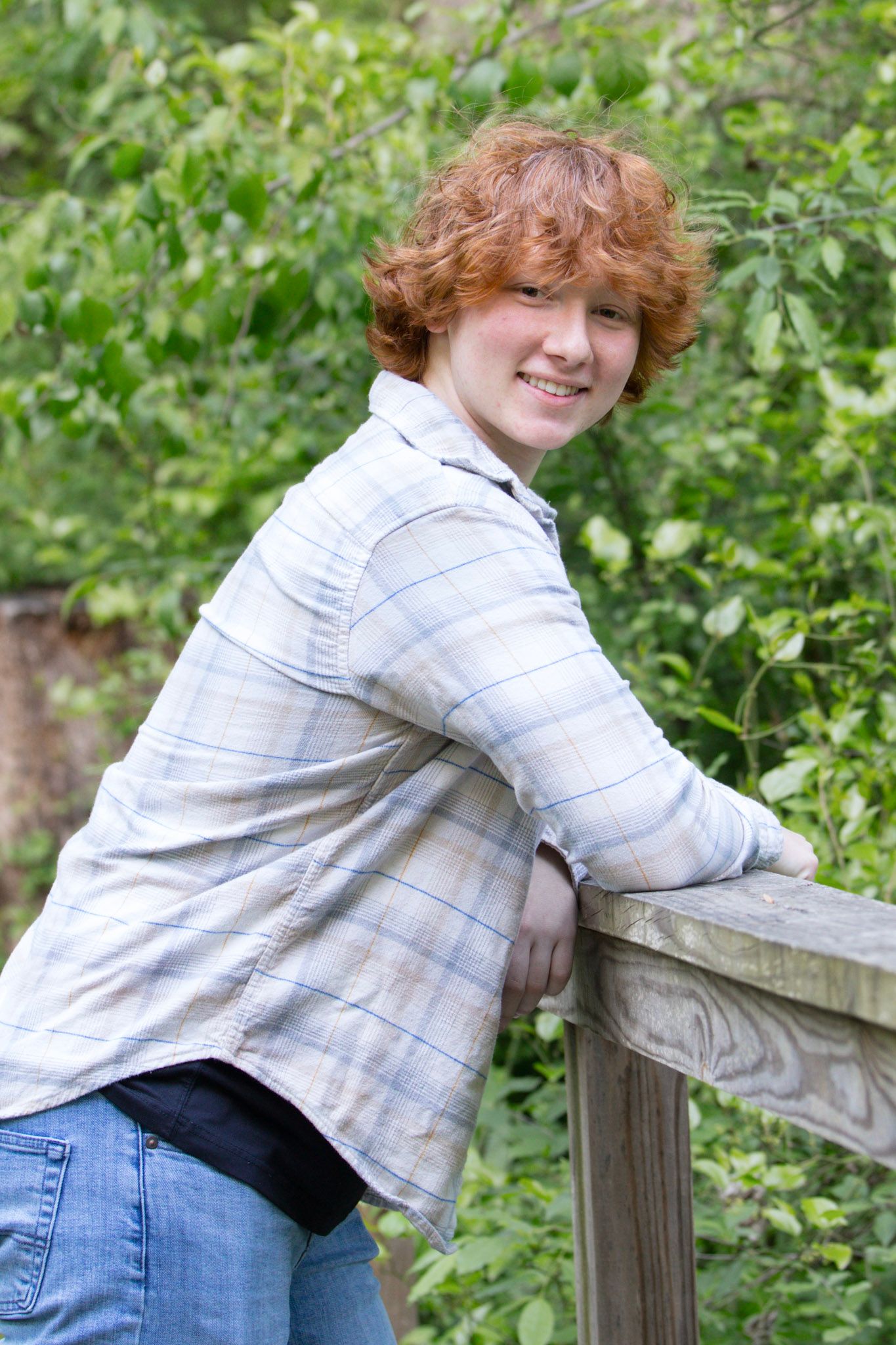 Smiling redhead teen leaning on wooden railing outdoors, wearing plaid shirt and jeans with green foliage background.