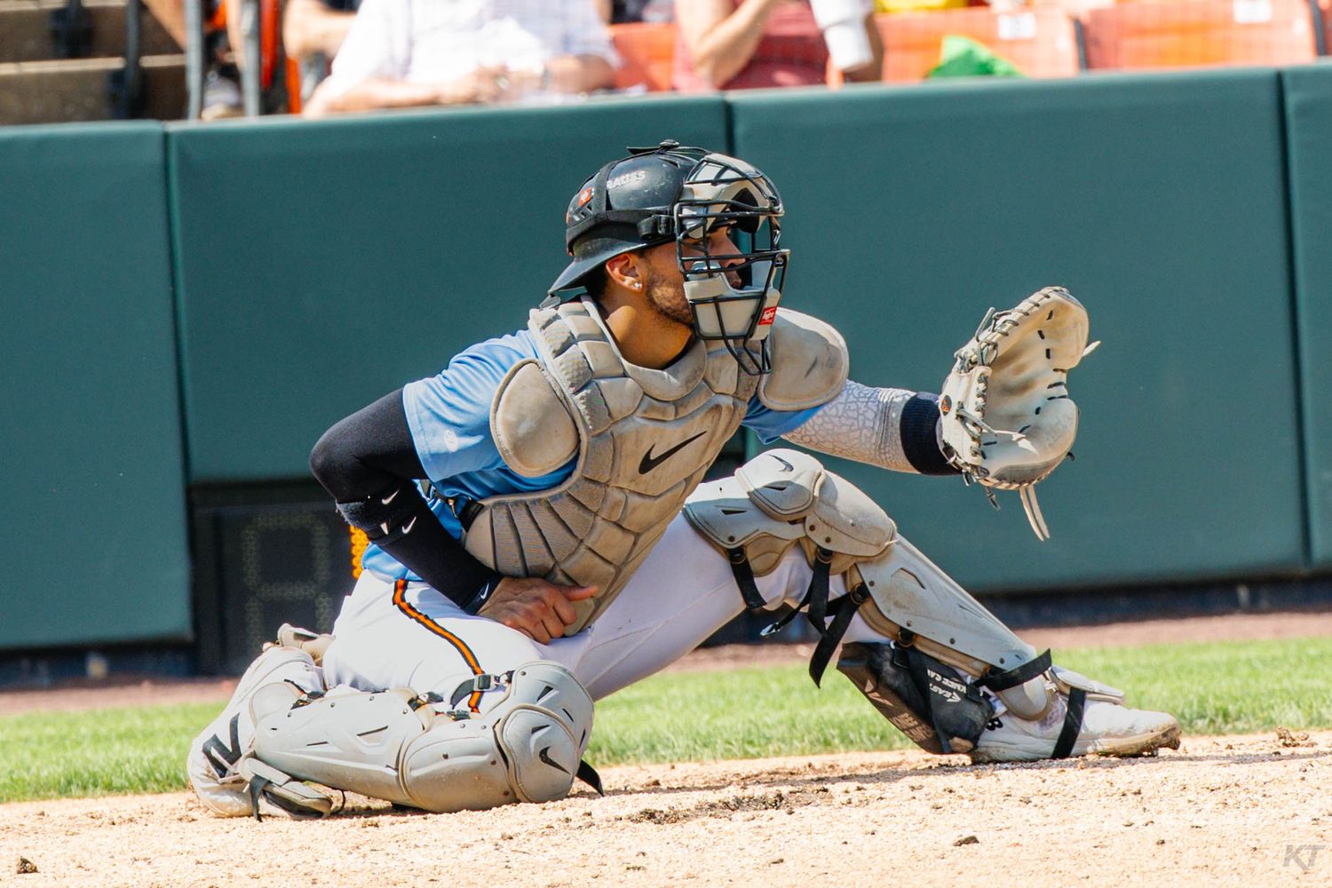 Baseball catcher in full protective gear crouching behind home plate, wearing helmet and mitt, ready to receive a pitch.