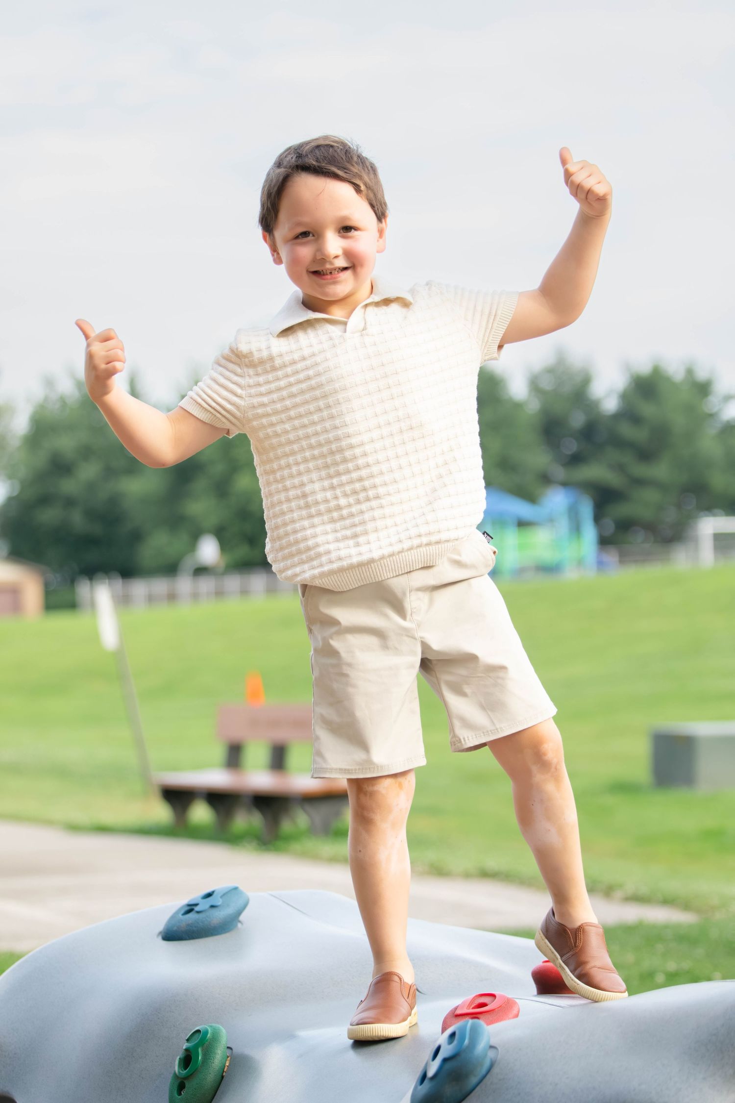 Happy young boy standing on playground climbing rock with thumbs up, smiling outdoors on sunny day.