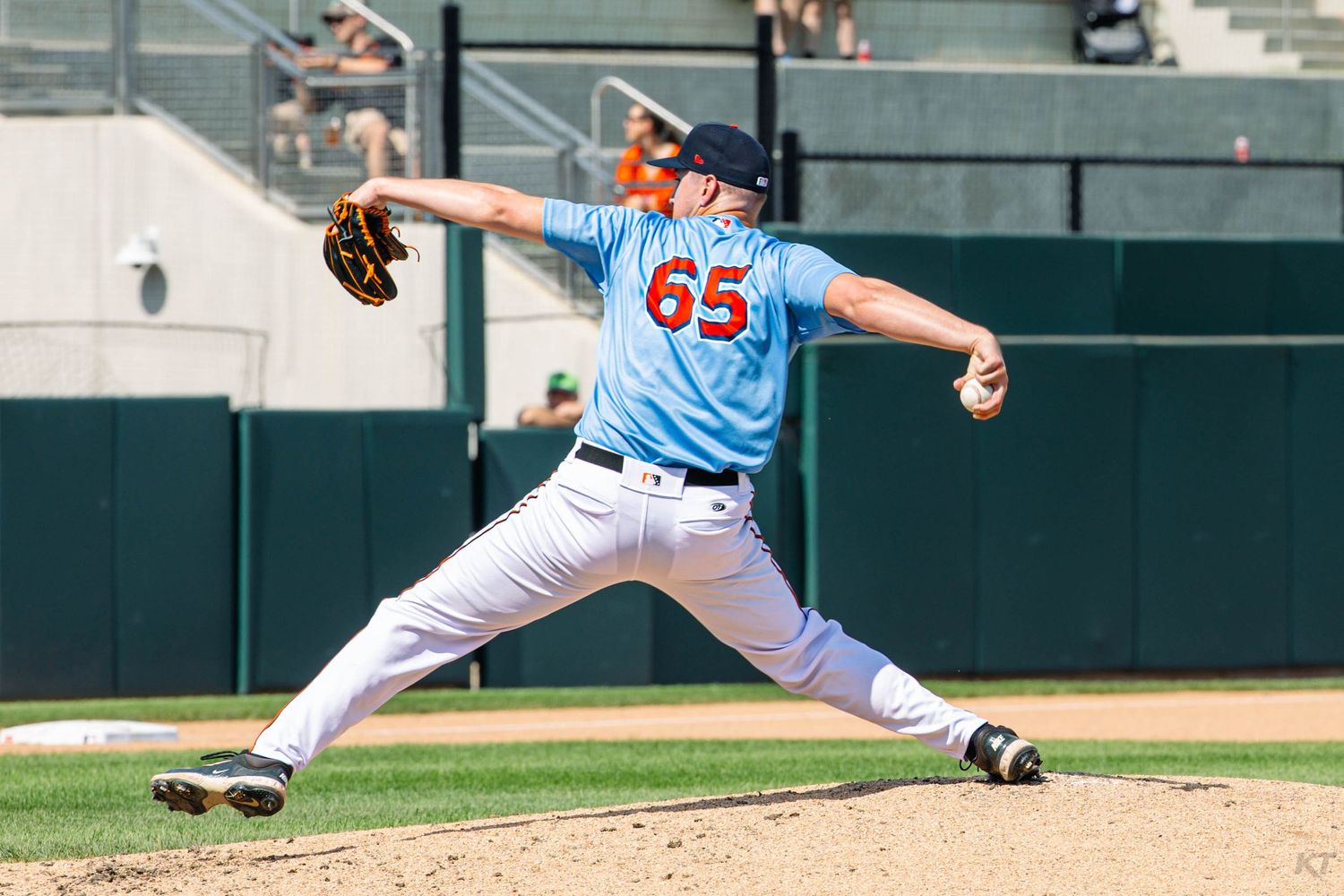 Baseball pitcher #65 in light blue uniform delivers a pitch during a sunny minor league game.