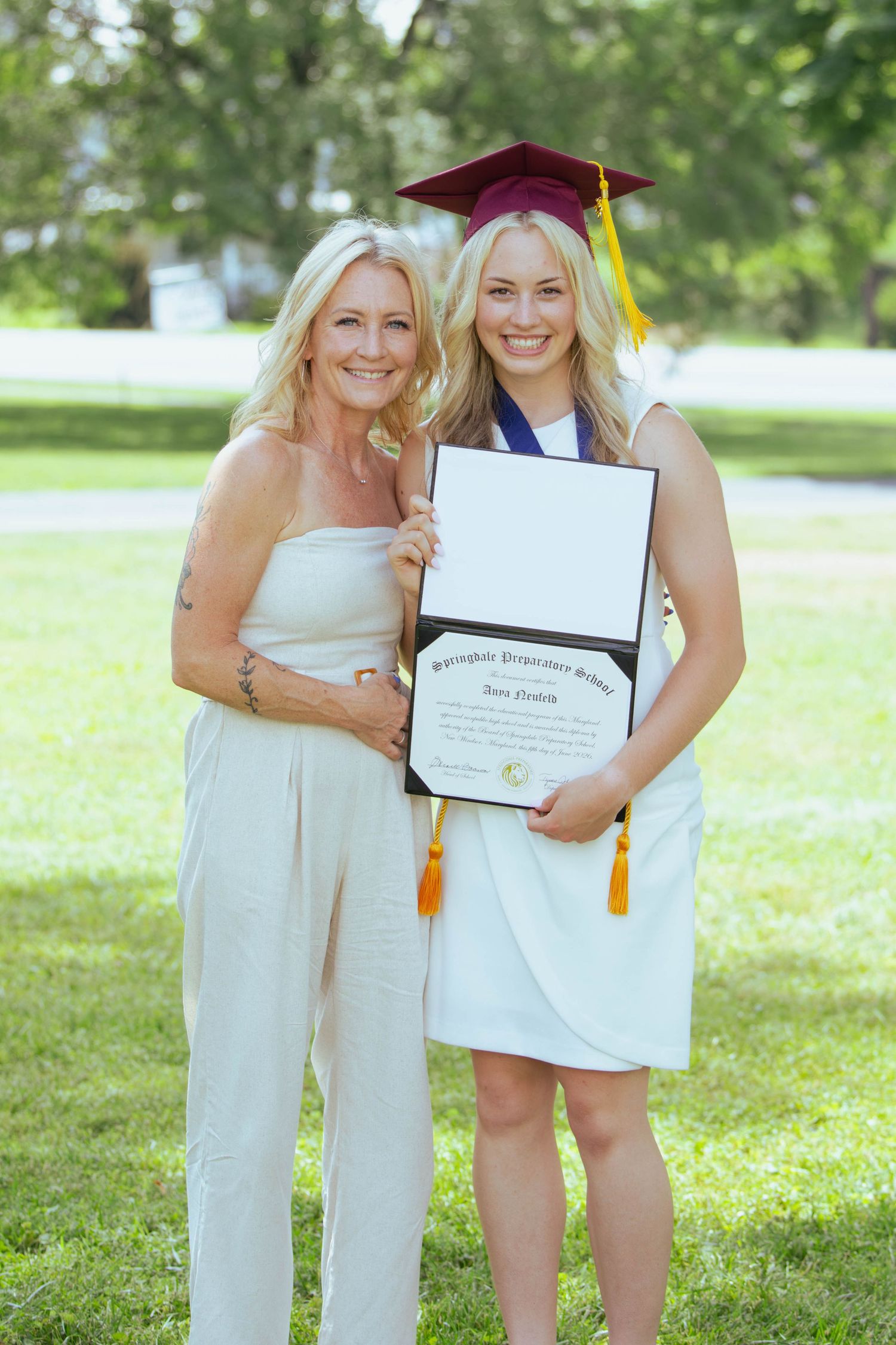 Two women in white outfits pose outdoors, one wearing a graduation cap and holding a diploma at a graduation ceremony.