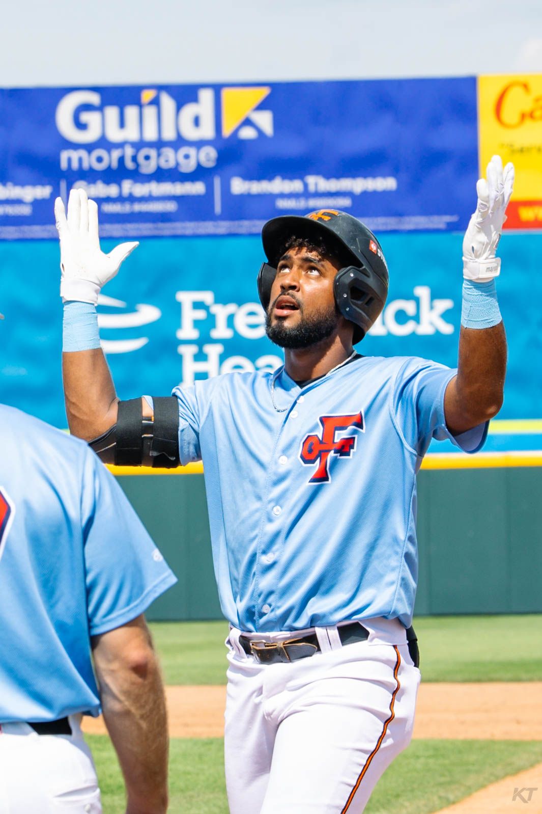 Baseball player in light blue uniform raises hands in celebration at a sunny stadium with Guild Mortgage signage.