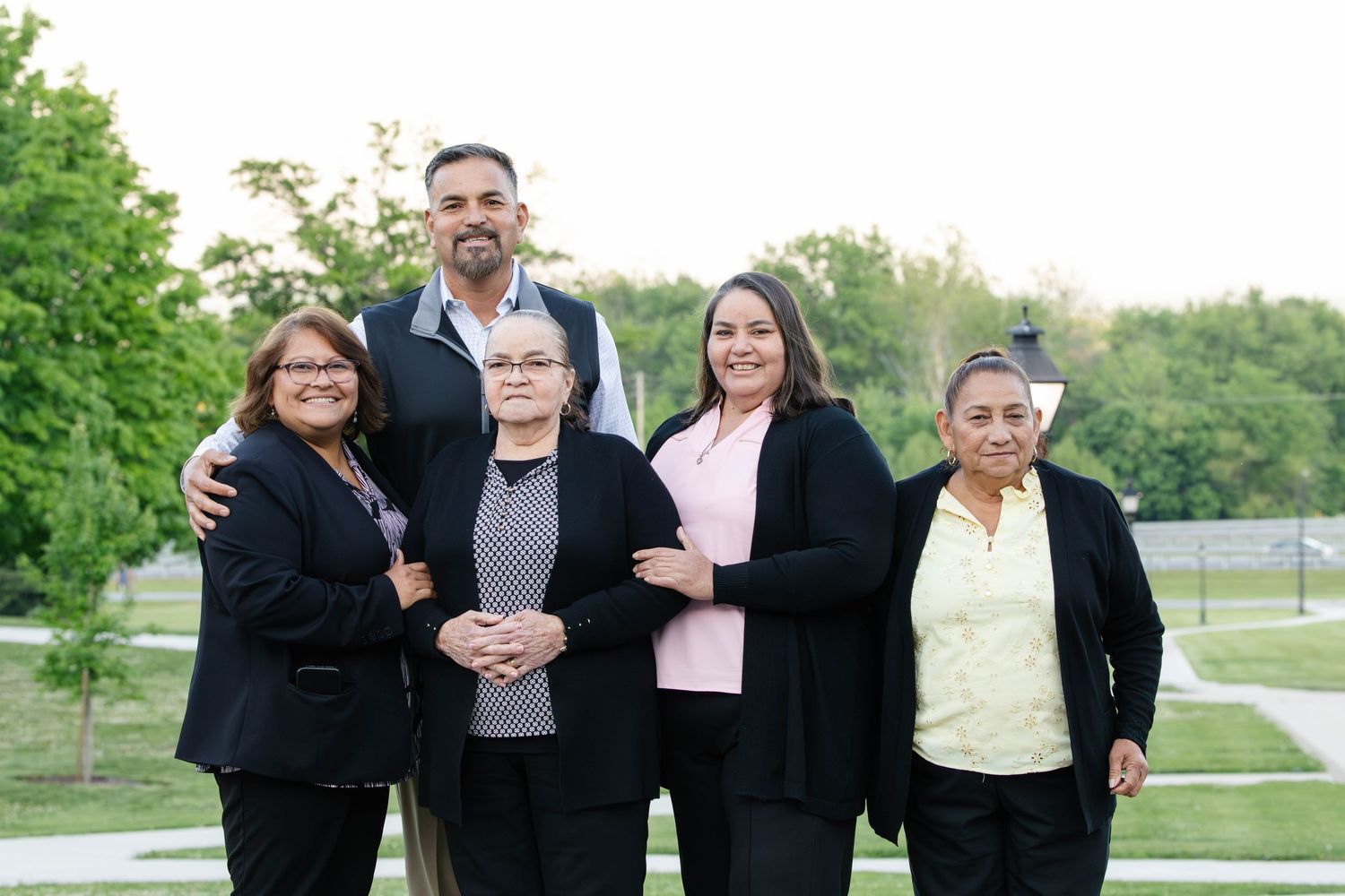 Group of five smiling adults posing outdoors in park setting.