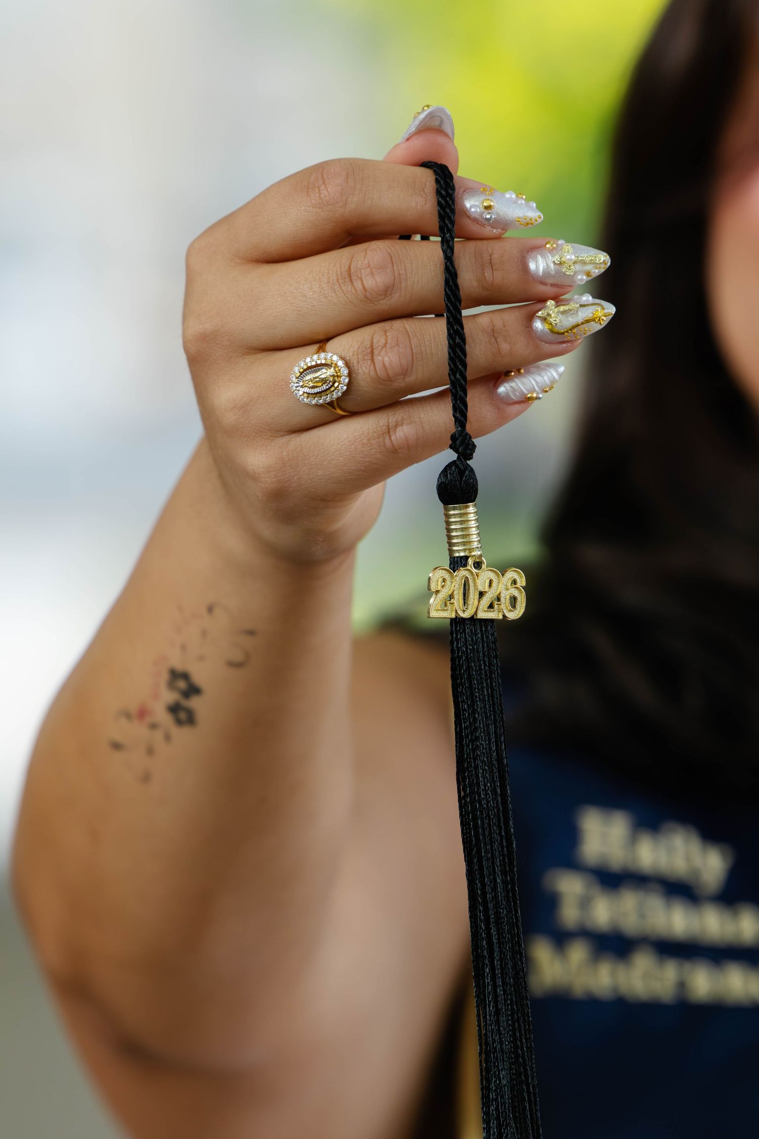 Close-up of graduate hand holding 2026 gold tassel charm with decorative acrylic nails and Virgen de Guadalupe ring.