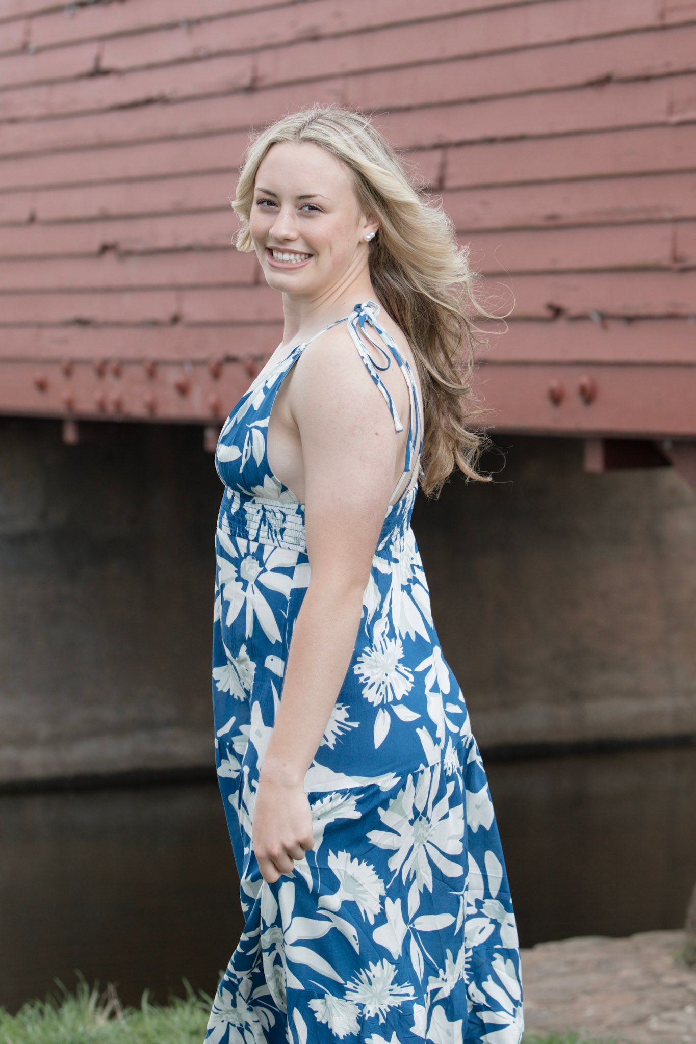 Smiling blonde woman in blue floral halter dress near red covered bridge.