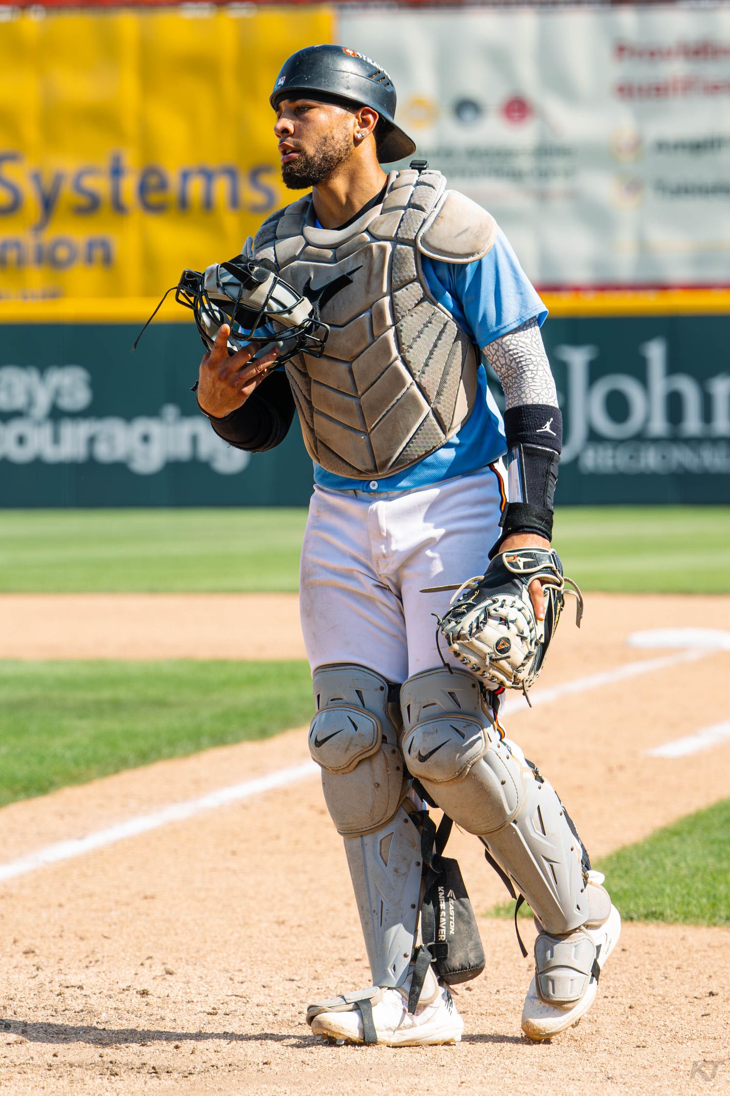 Minor league baseball catcher in full protective gear walks on field during a sunny afternoon game.