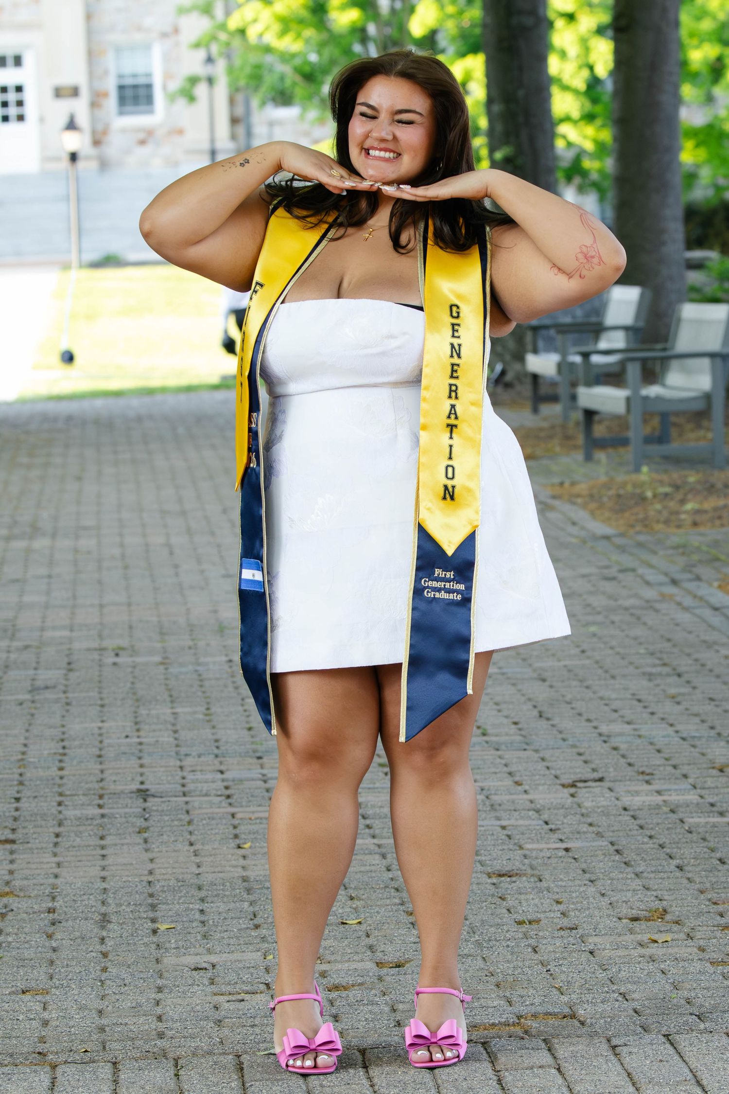 Happy female graduate posing outdoors wearing yellow GENERATION stole and First Generation Graduate blue sash with pink heels.