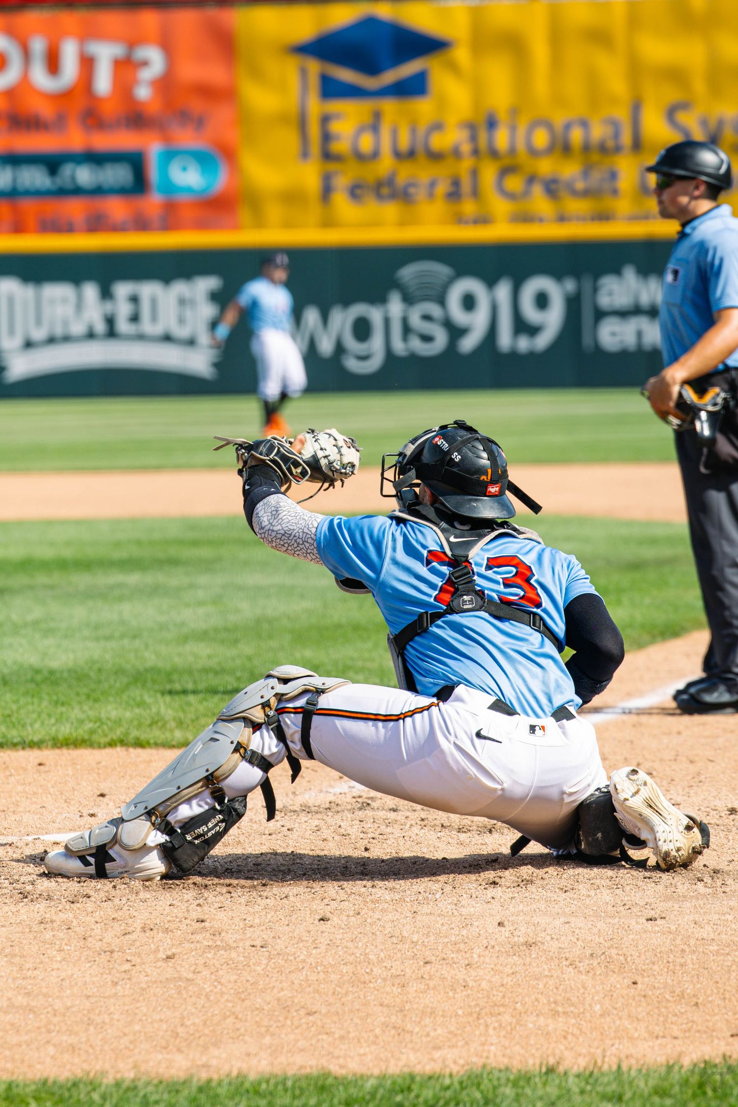 Baseball catcher in blue jersey crouching to catch a pitch during a sunny game, with an umpire standing behind.