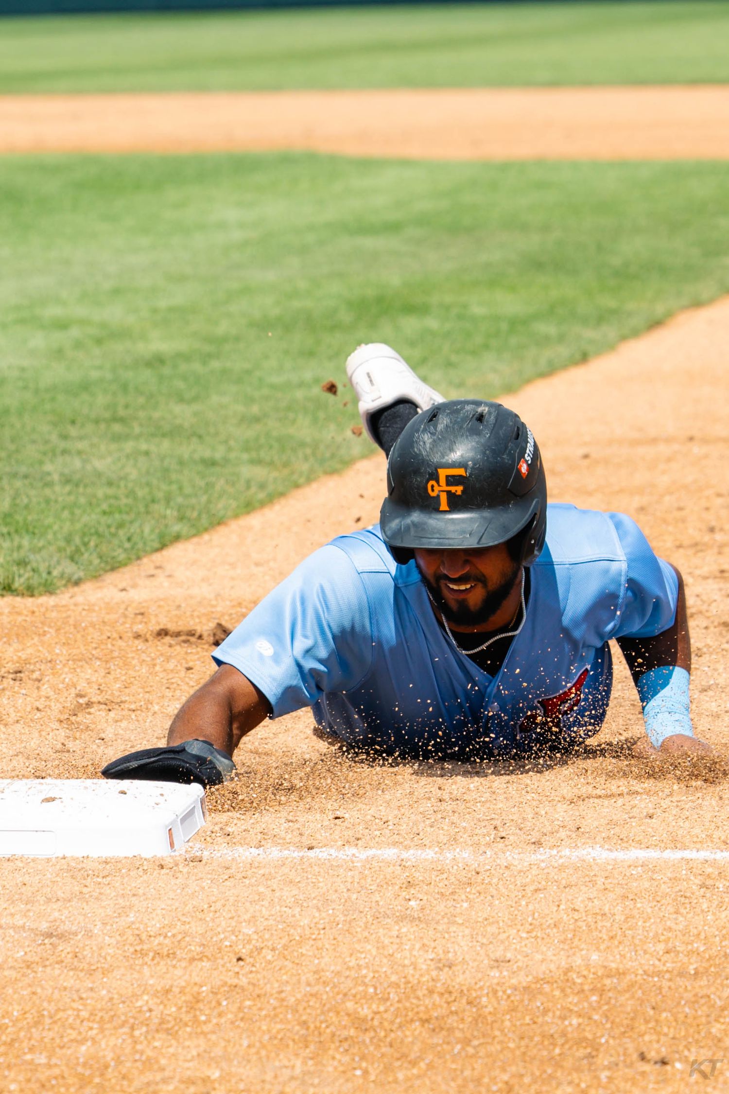 Baseball player in blue uniform diving headfirst into base, sending dirt flying on sunny day.
