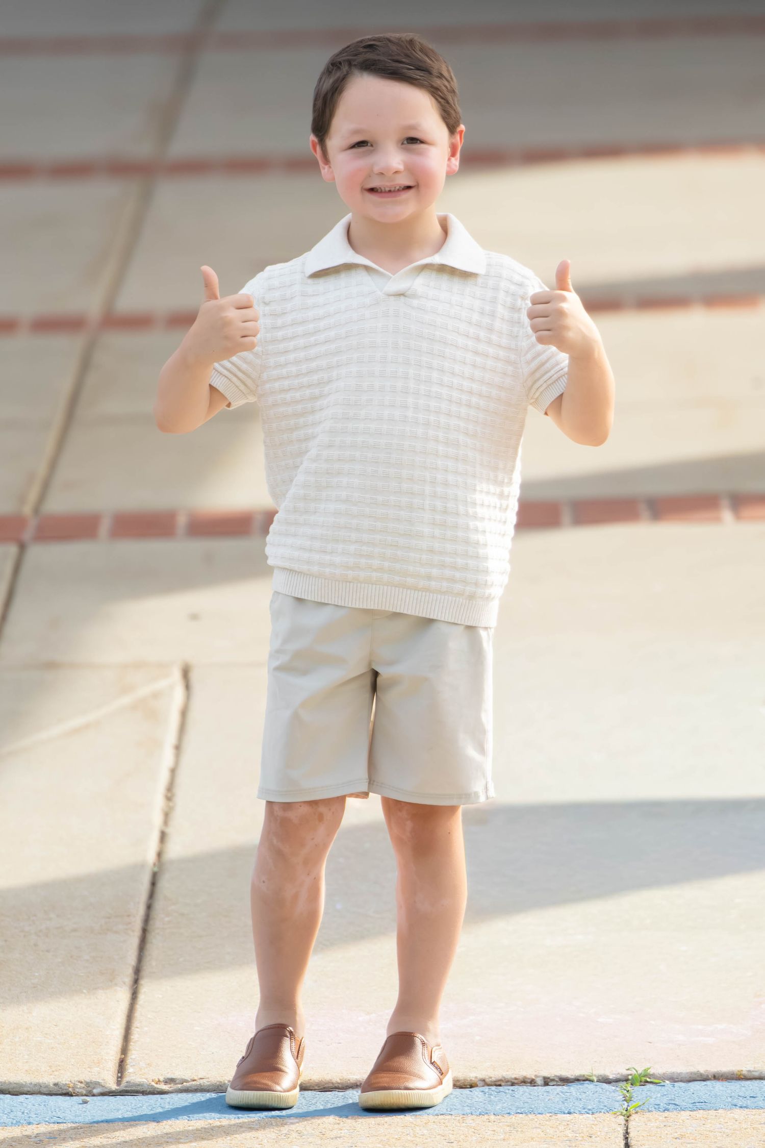 A cheerful young boy in a white polo shirt and khaki shorts giving two thumbs up outdoors.
