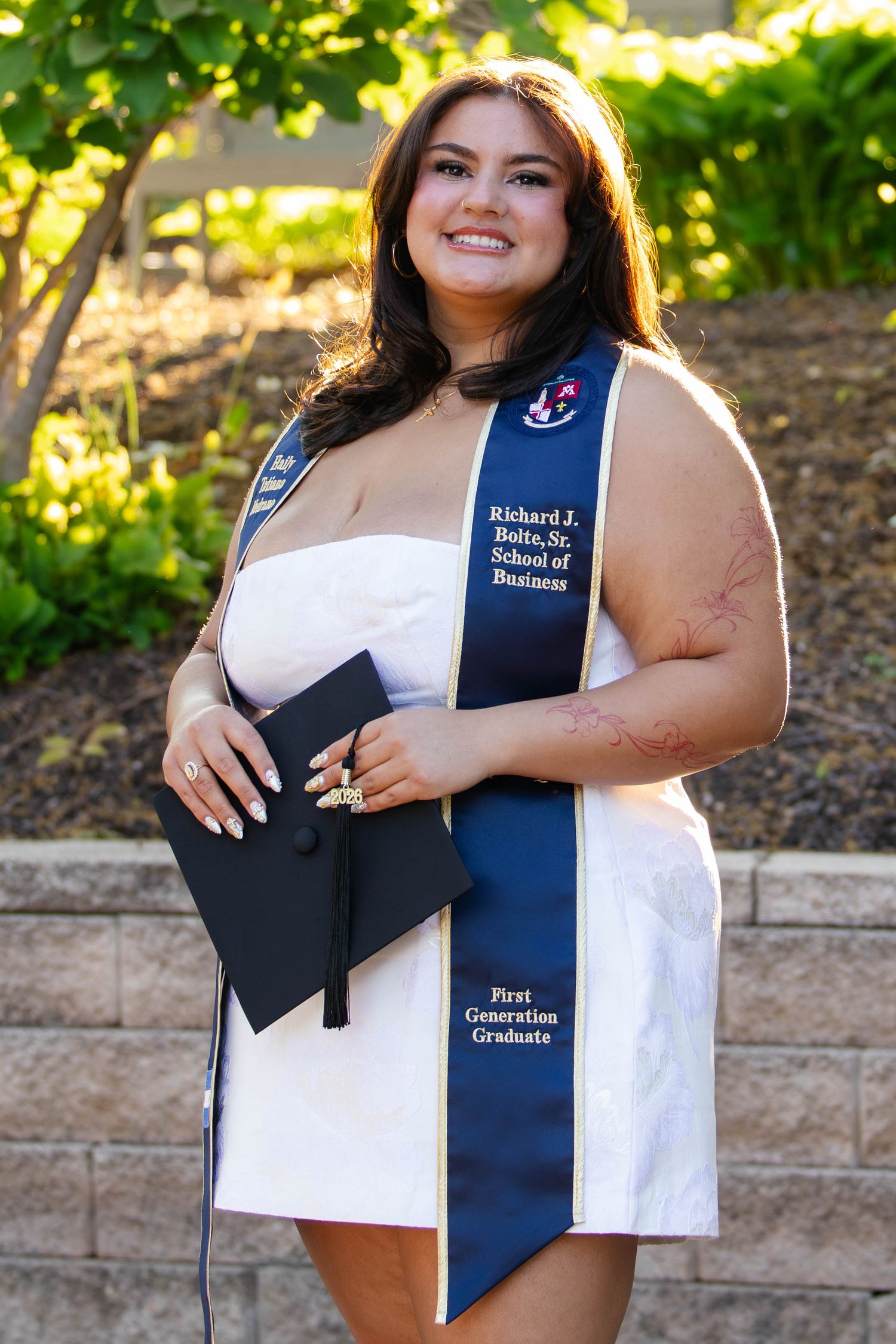 Smiling female graduate wearing white dress and navy blue graduation stole from Richard J. Bolte Sr. School of Business, holding cap.