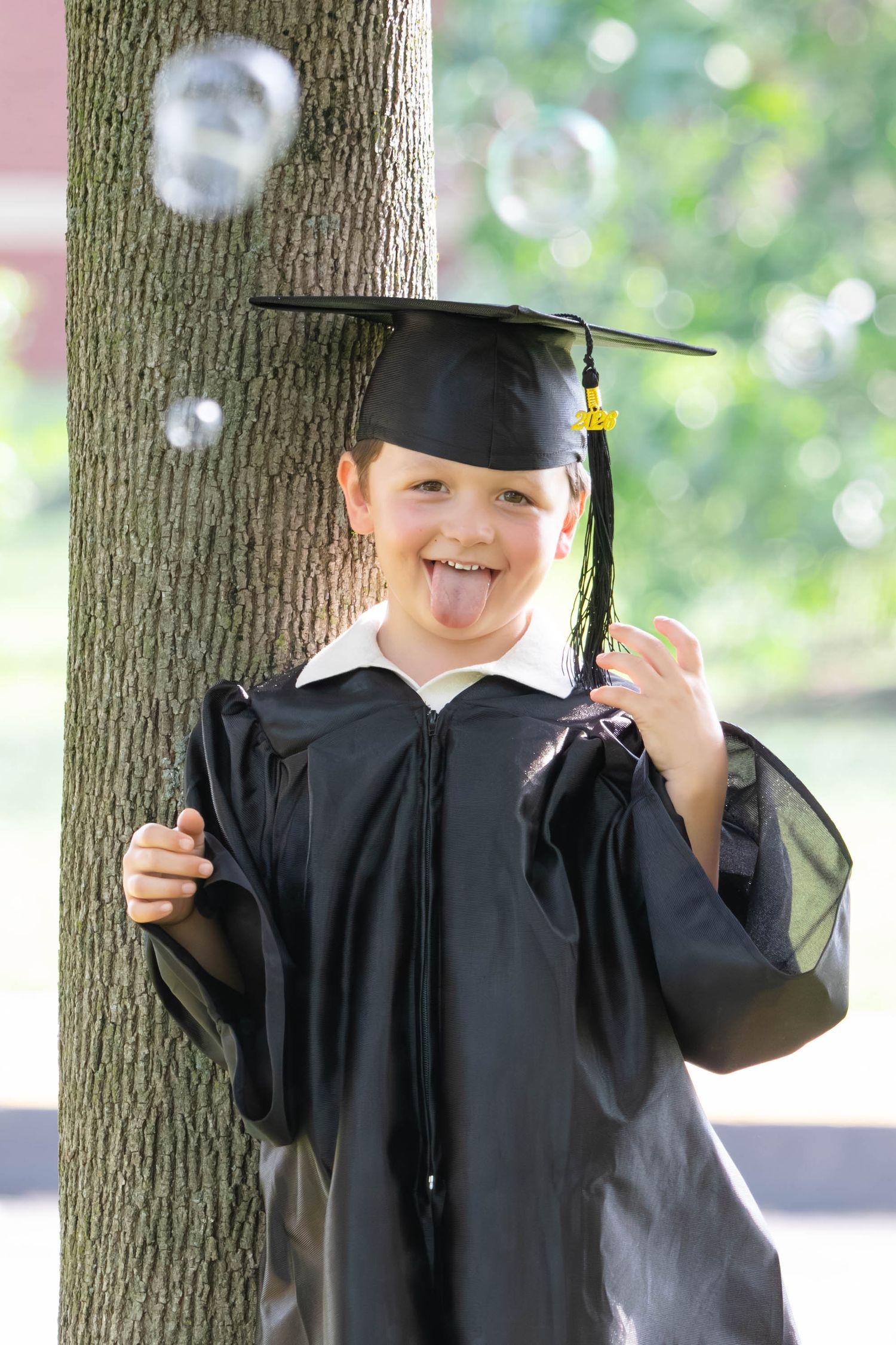 Young boy in black graduation cap and gown sticking tongue out while leaning against a tree outdoors.