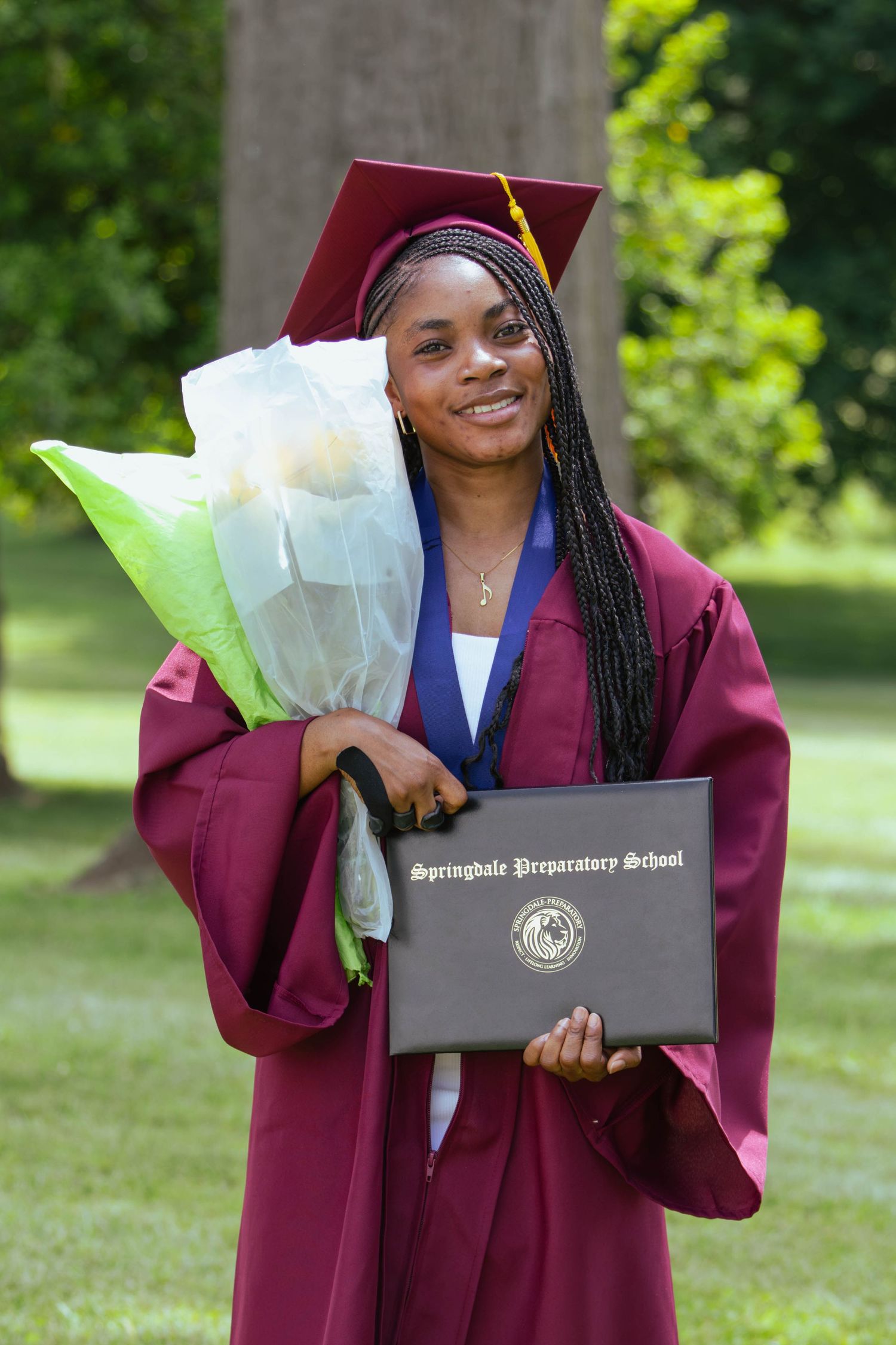 Young woman in maroon graduation cap and gown smiles holding diploma from Springdale Preparatory School and bouquet outdoors.