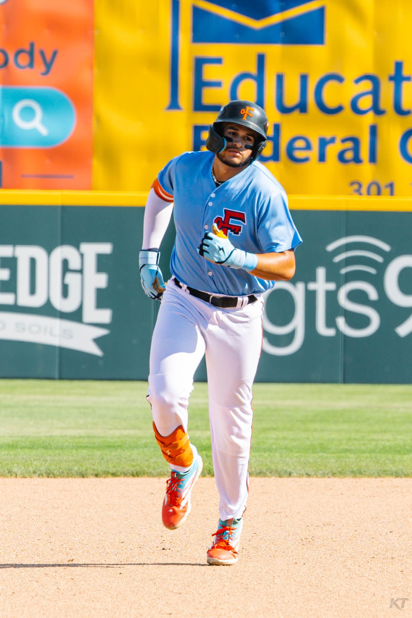 Baseball player in blue and orange uniform running bases during a sunny minor league game.