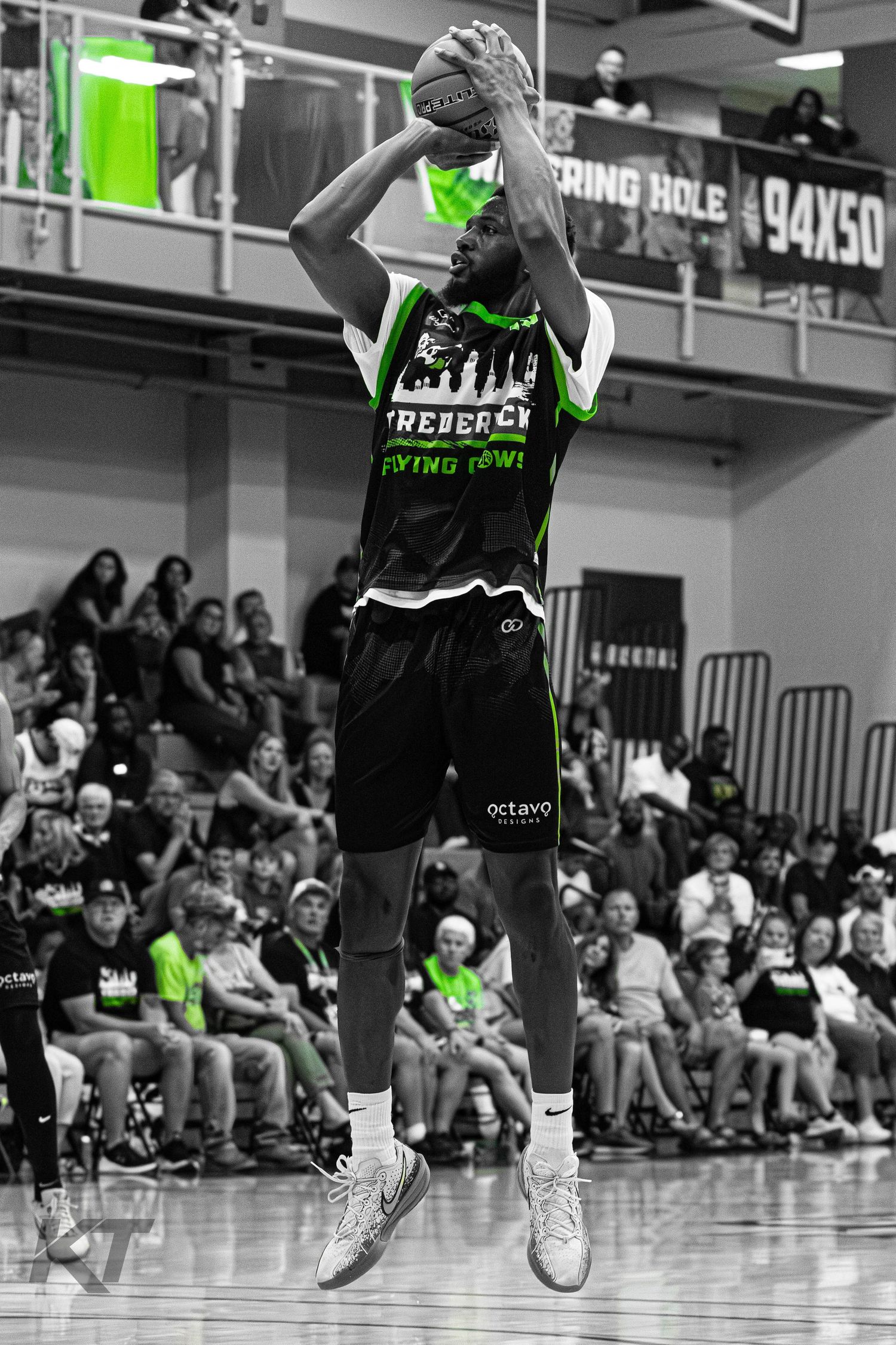 Basketball player in green and black uniform jumps to shoot during a game, crowd watching in black and white background.