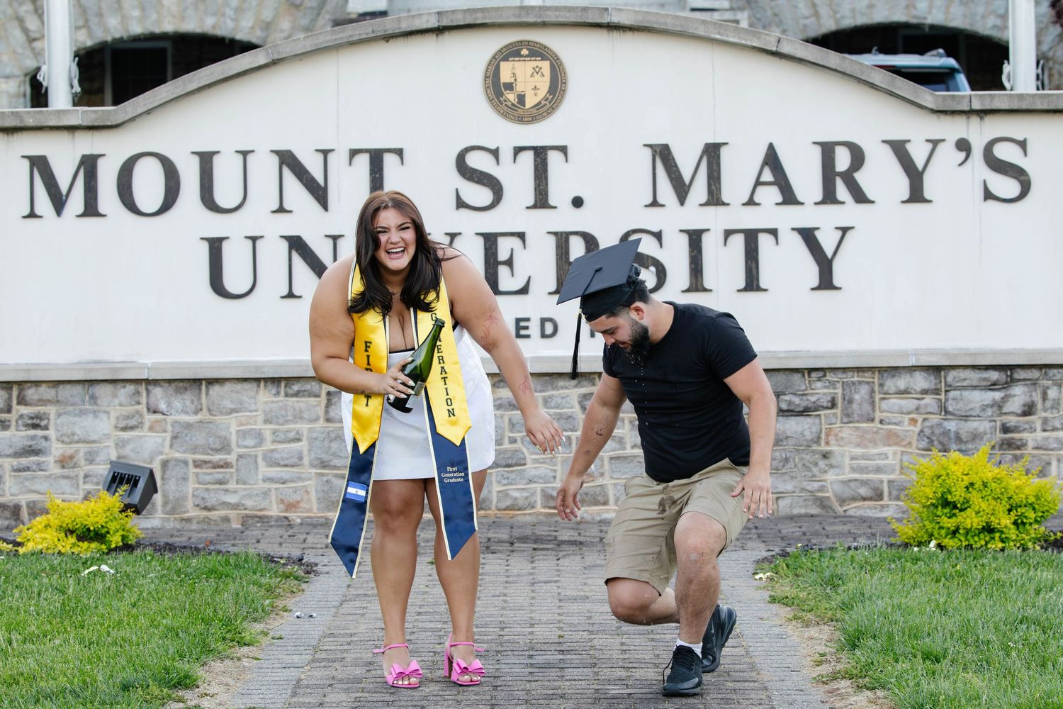 Female graduate in yellow sash poses with man outside Mount St. Mary's University entrance sign.