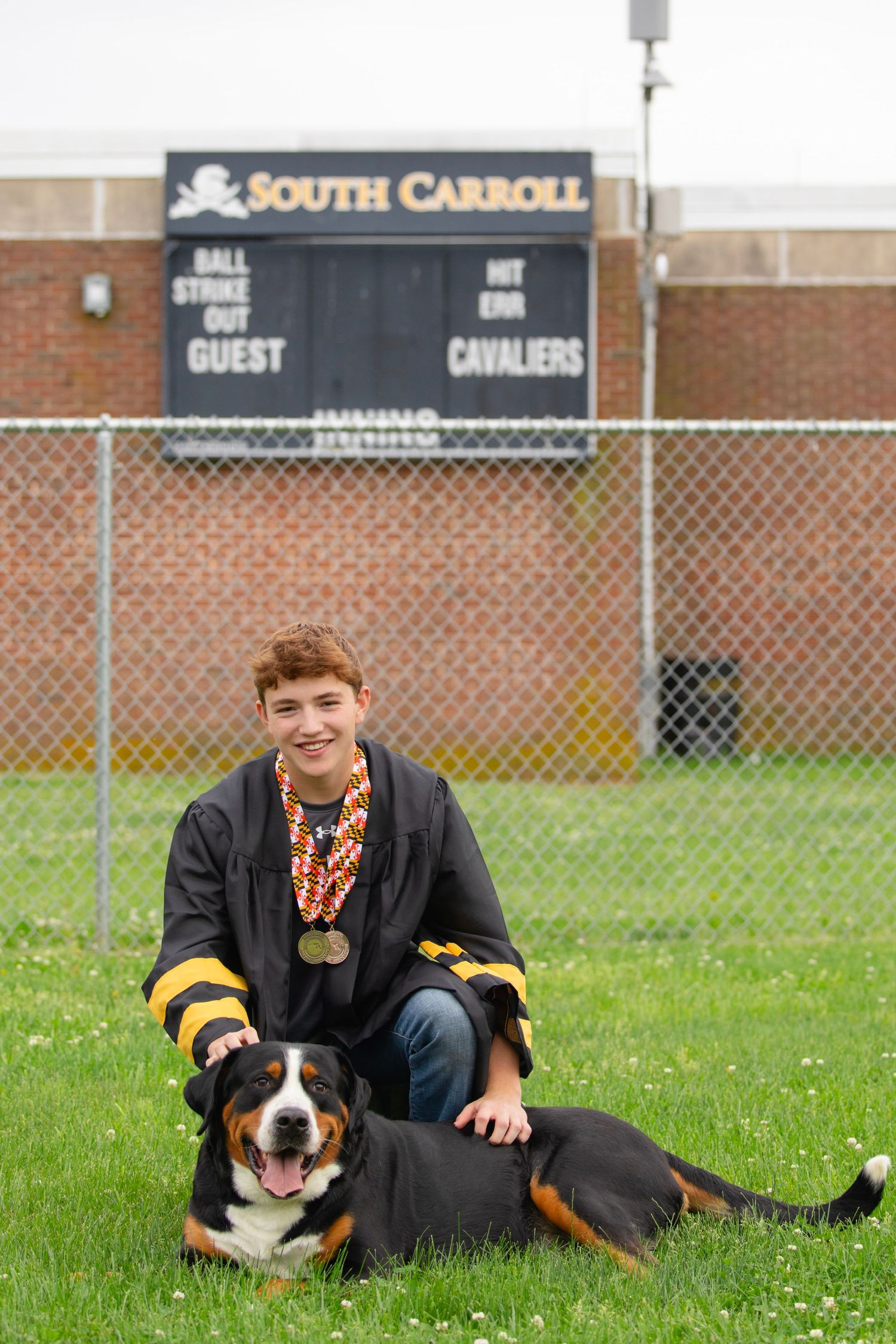 A smiling graduate in black gown with medals poses with a Bernese Mountain Dog at South Carroll Cavaliers field.