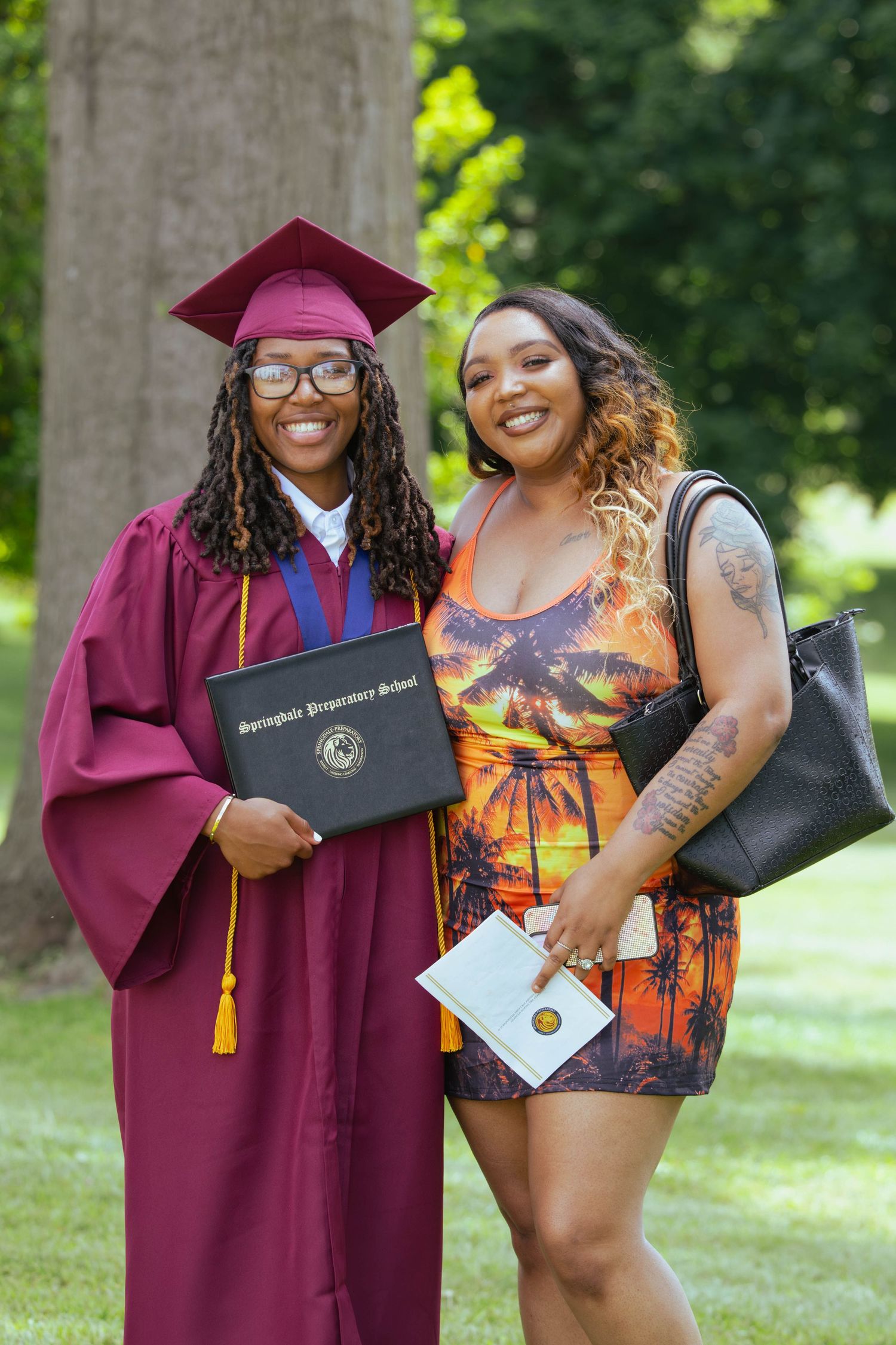Two women posing outdoors, one wearing maroon graduation cap and gown holding diploma at Springdale Preparatory School.