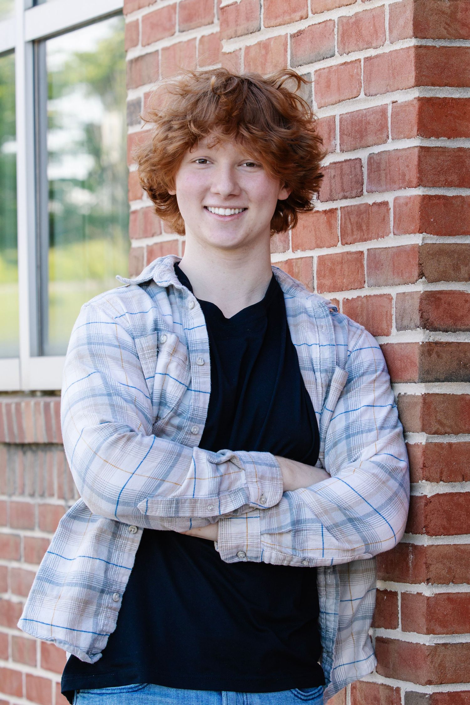 Smiling teenage boy with curly red hair leaning against brick wall, wearing plaid shirt over black tee.