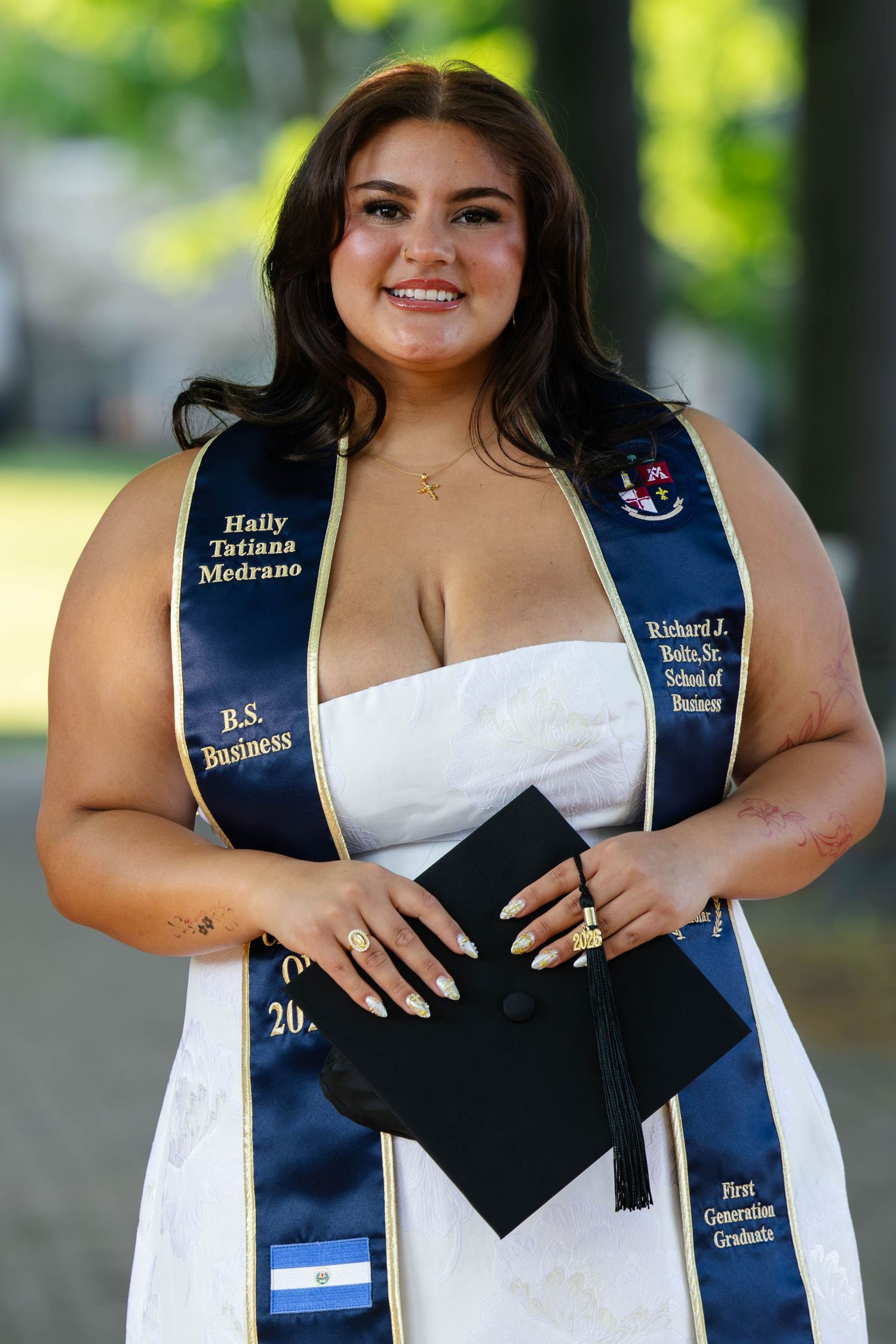 Graduate Haily Tatiana Medrano in white dress with B.S. Business sash holding cap.
