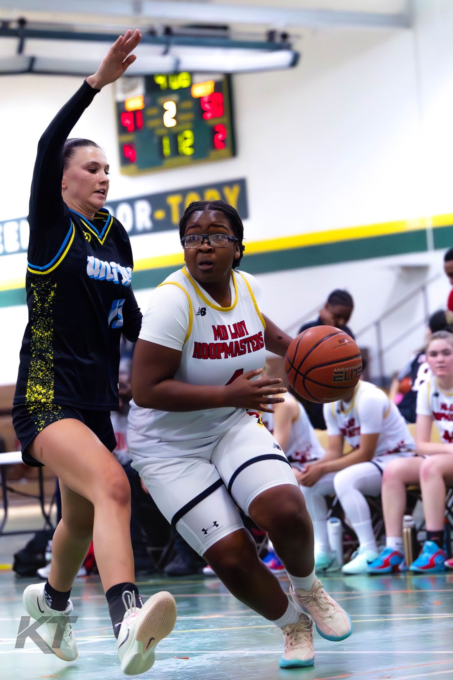 Female basketball player in white uniform drives to the basket against defender in black during indoor game.