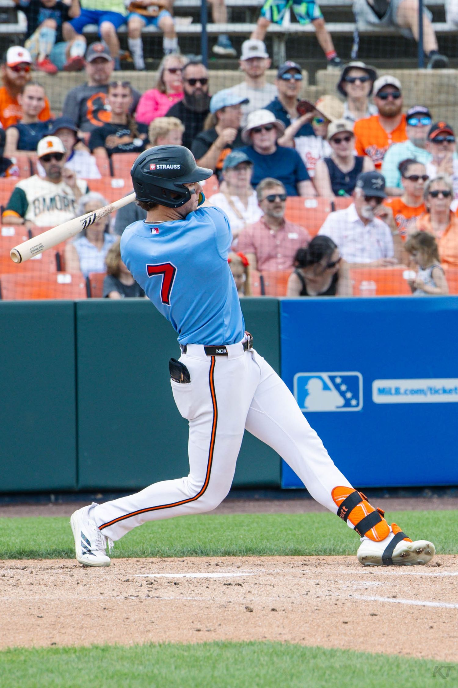 Baseball player #7 swings bat in light blue uniform at minor league game with fans in orange and navy in background.