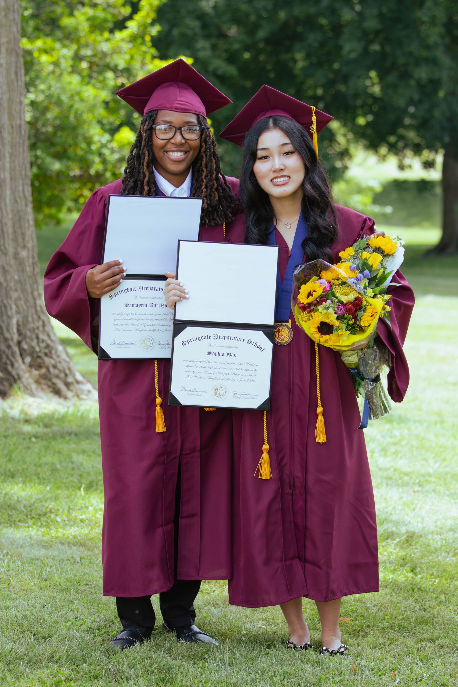 Two graduates in maroon caps and gowns proudly hold diplomas and flowers outdoors on a sunny day.