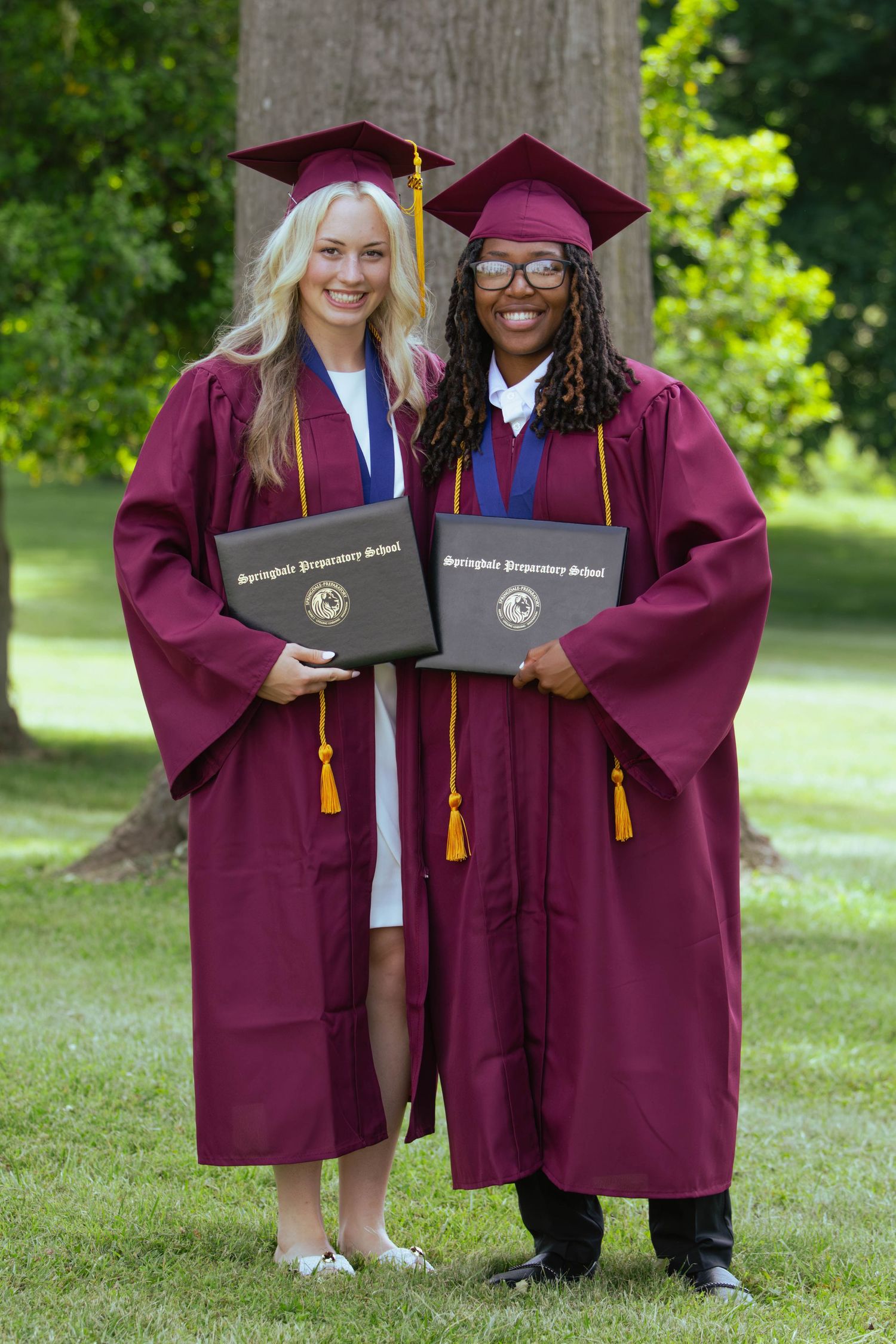 Two graduates in maroon cap and gown hold diplomas outdoors at Springdale Preparatory School graduation ceremony.