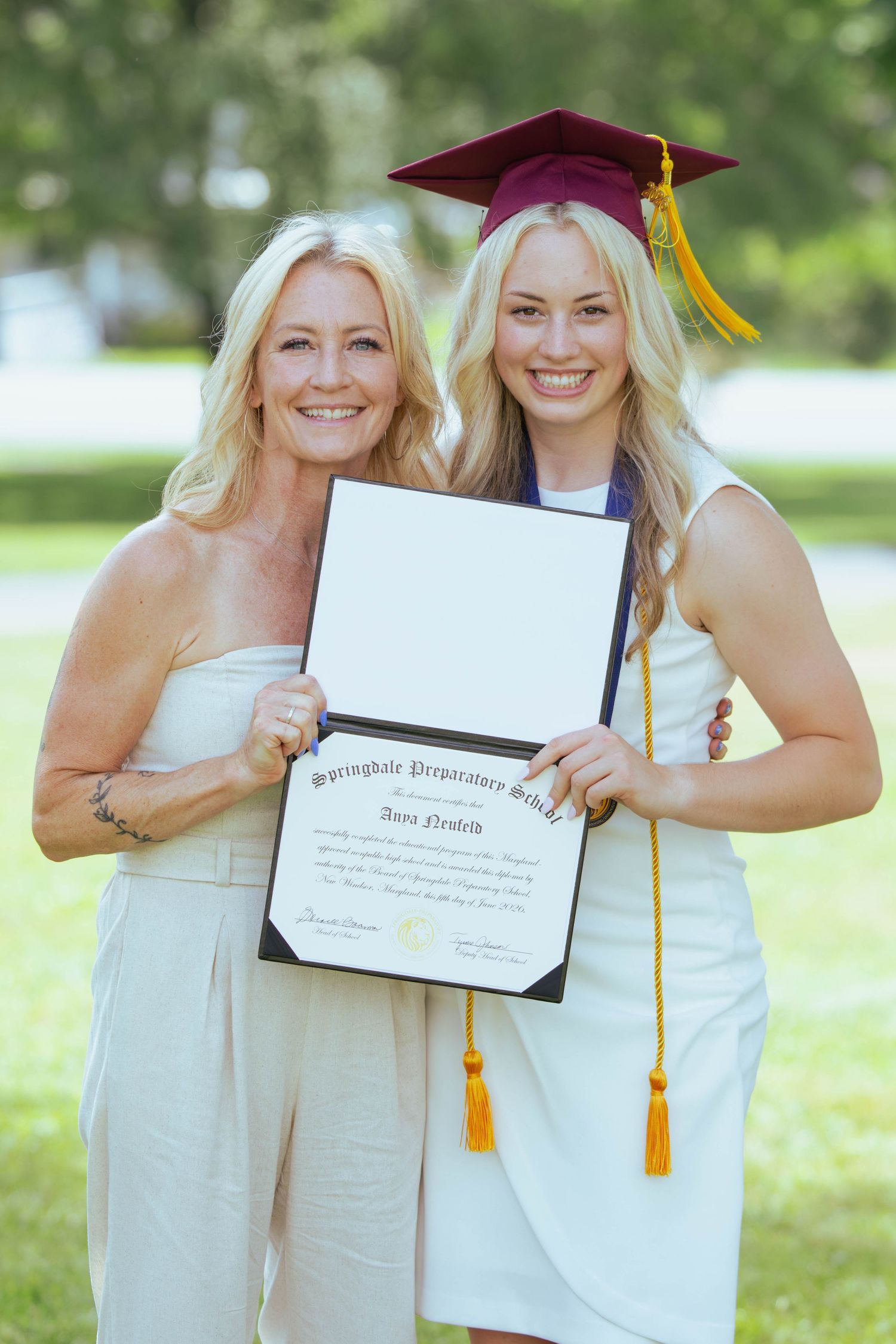 Two blonde women proudly display a graduation diploma outdoors, one wearing a maroon cap and gown with gold tassel.