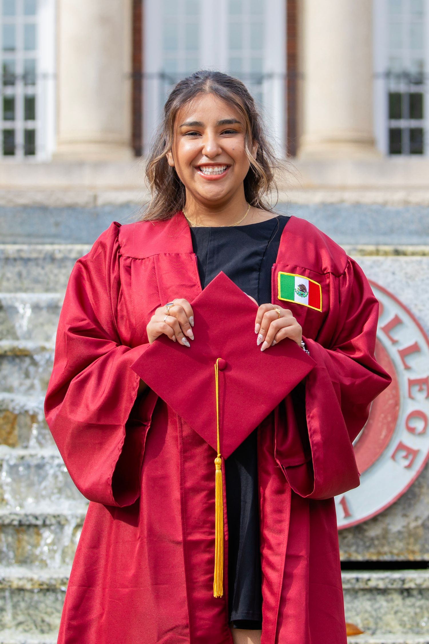 Smiling graduate in red cap and gown holding mortarboard with Mexican flag patch.